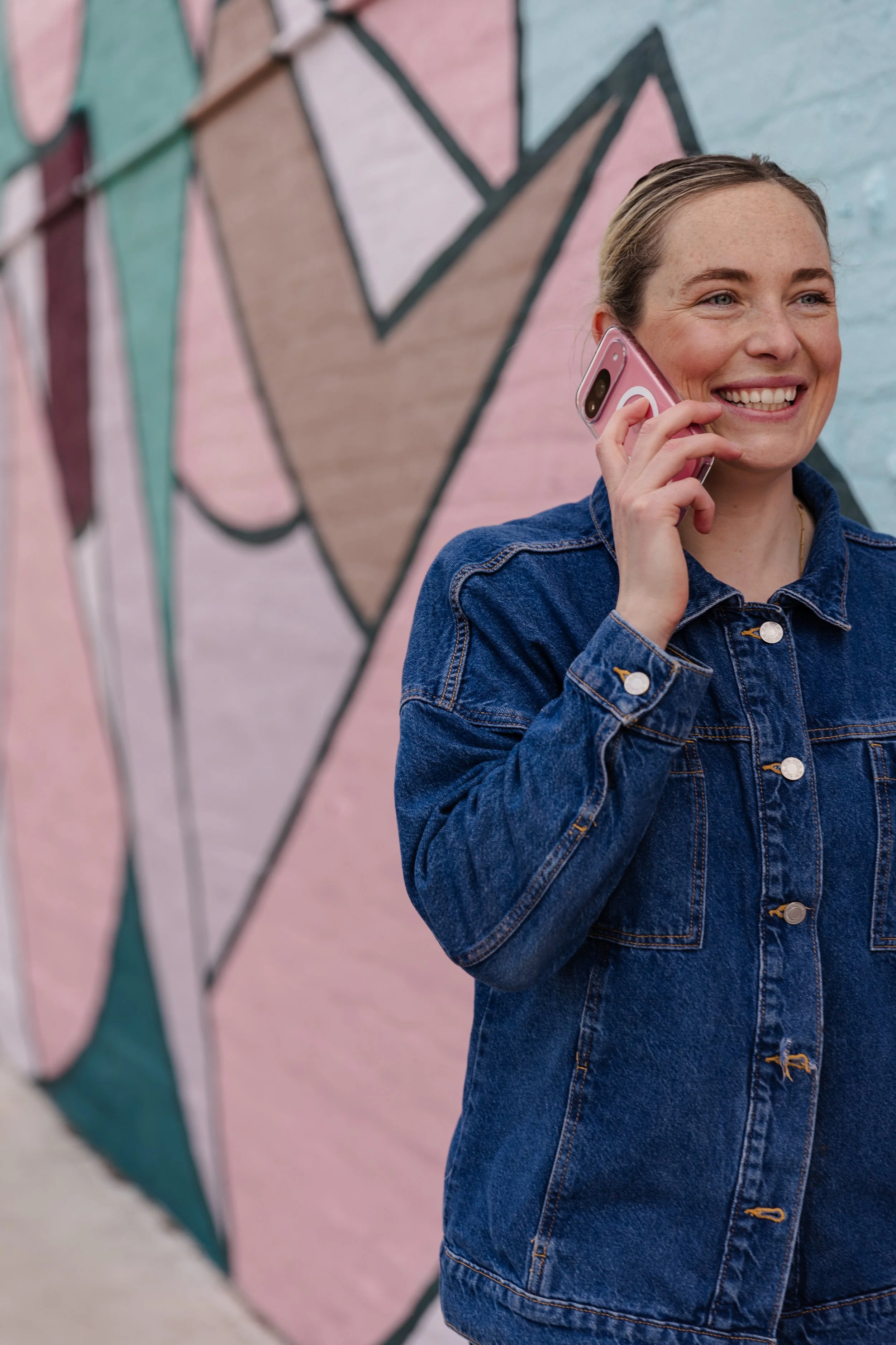Imogen, a woman, wearing a denim jacket talking on a pink cell phone, standing in front of a colorful geometric mural on a brick wall.