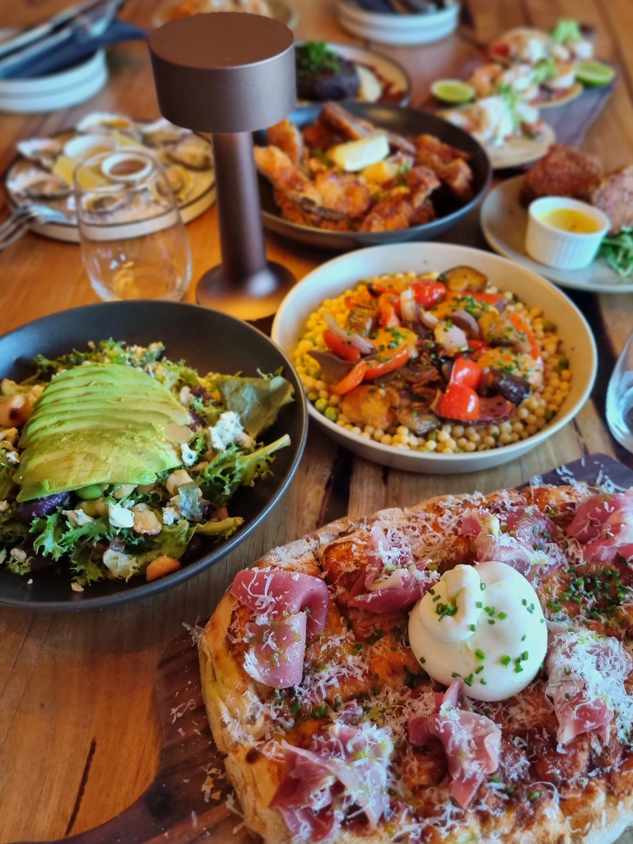A spread of assorted Italian and Mediterranean dishes including pizza, salad with avocado, couscous with vegetables, fried chicken, oysters, and other small plates on a wooden table.
