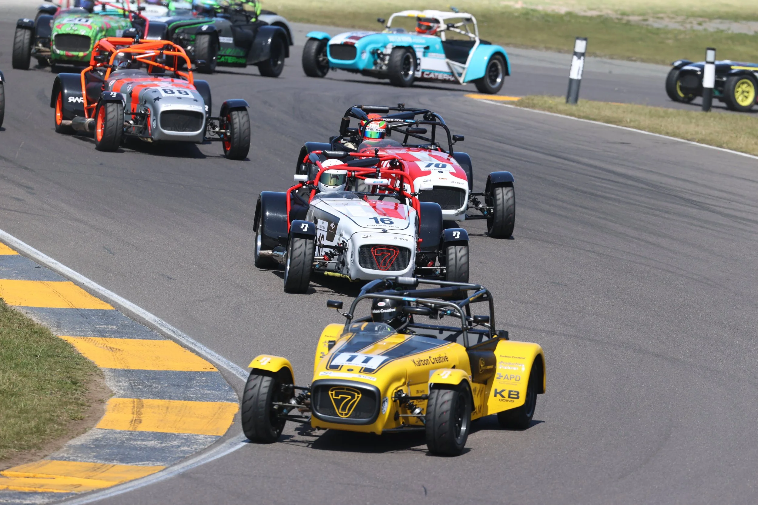 Several open-wheel race cars in red, yellow, white, and blue driving on a race track during a race.