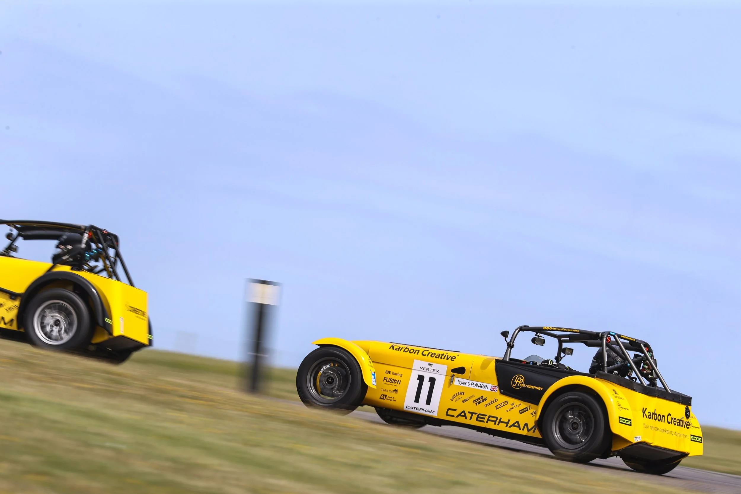 Two yellow Caterham race cars speeding on a track with a blue sky background.