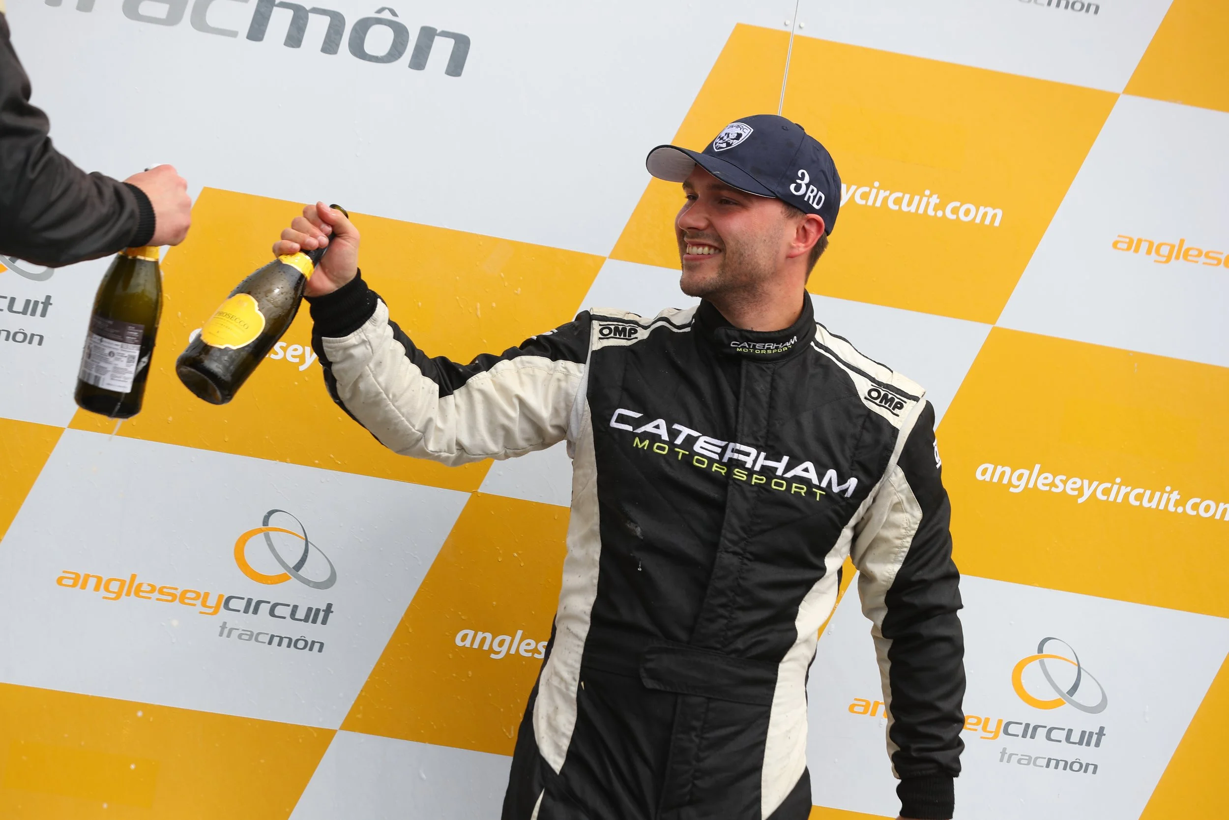 Race car driver in a black and white racing suit celebrating with champagne on a podium at an race event at Anglesey Circuit.