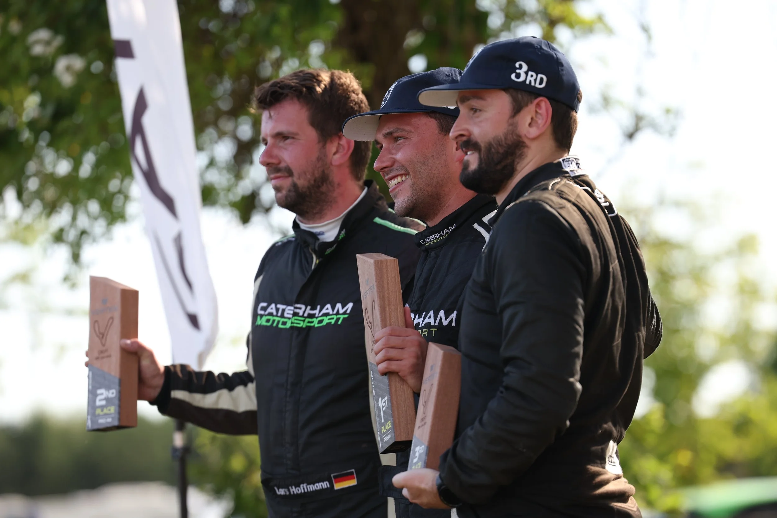 Three men in racing suits holding trophies on a podium, celebrating at an outdoor motorsport event.