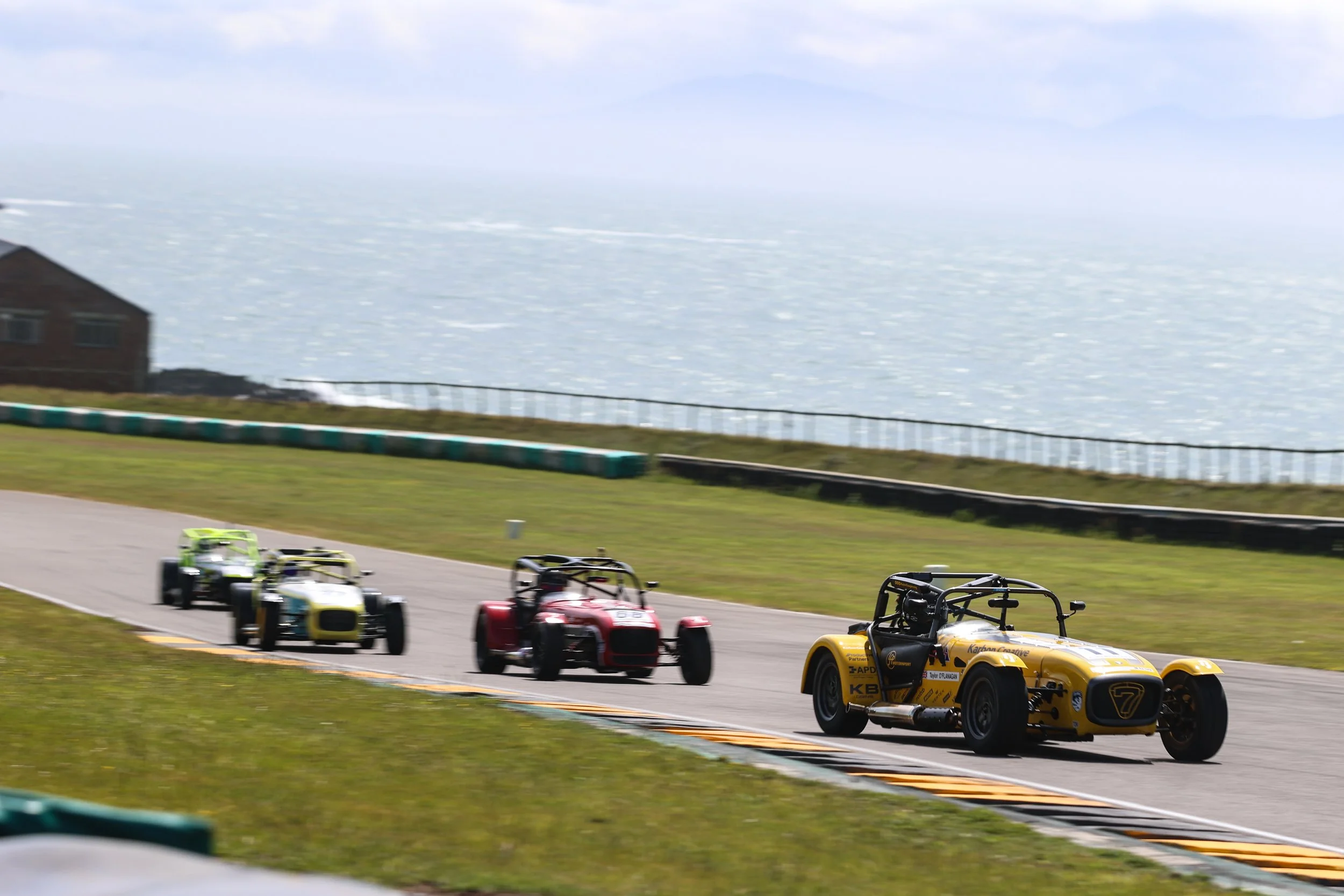 Four vintage race cars driving on a track near the water with a house on the grassy area to the side.