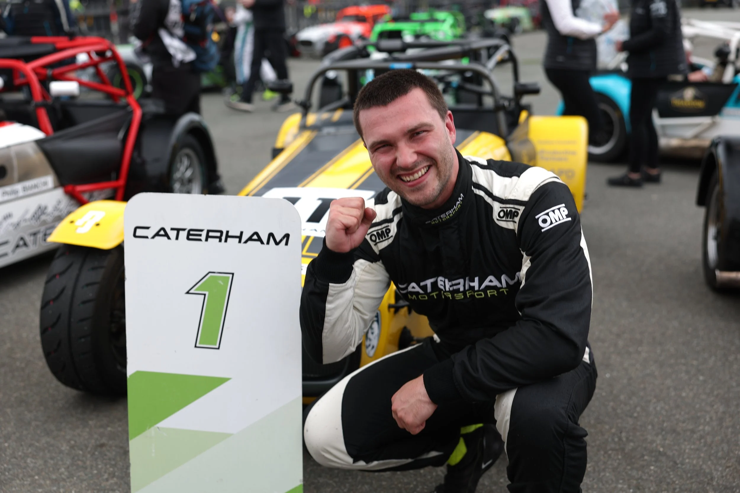 Race car driver celebrating first place victory, kneeling next to a sign that reads 'Caterham 1' with a racing car in the background.