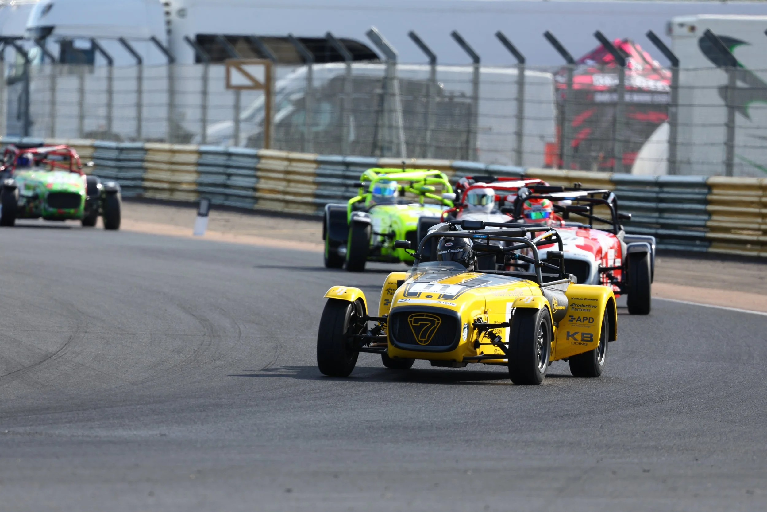 Four race cars on a track during a race, with the closest car being yellow with the number 7, followed by three other cars in various colors, navigating a curve.