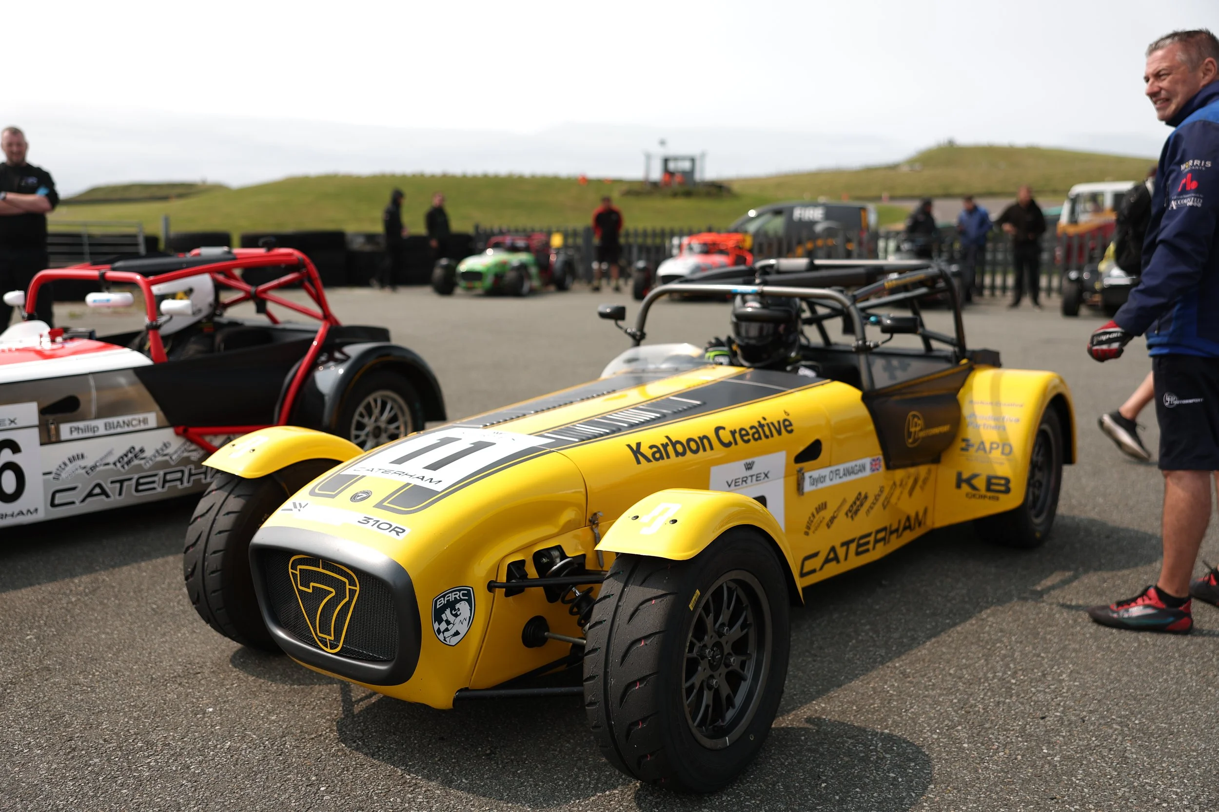 A yellow vintage race car with the number 7 on it, sponsored by Karbon Creative, parked at a racing event with other cars and people around.