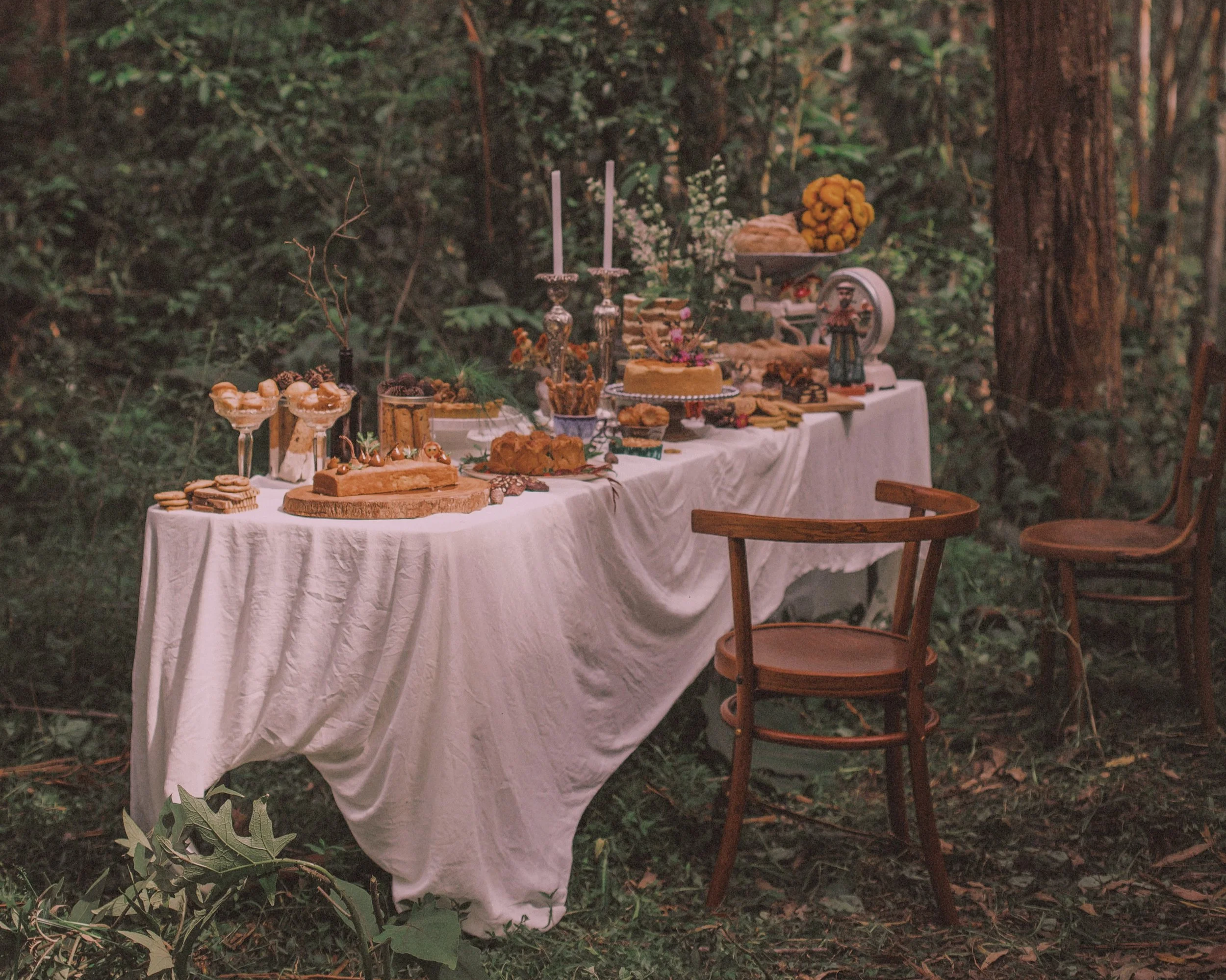 long table in the forest white tablecloth and stemmed glasses, cakes on pedestals, candles in silver candlesticks, breads and sweets inviting all into the feasting.