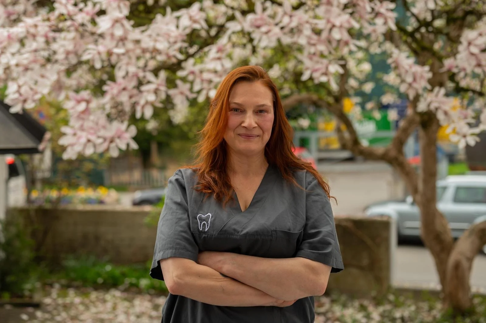 Frau mit roten Haaren in grauer Arbeitskleidung steht vor einem blühenden Baum im Freien.