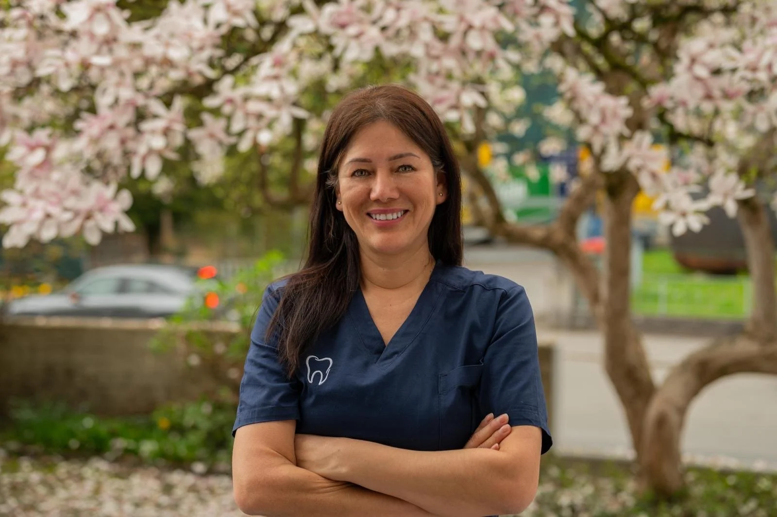 Frau in dunkelblauer Zahnarztuniform steht lächelnd vor blühenden Bäumen.