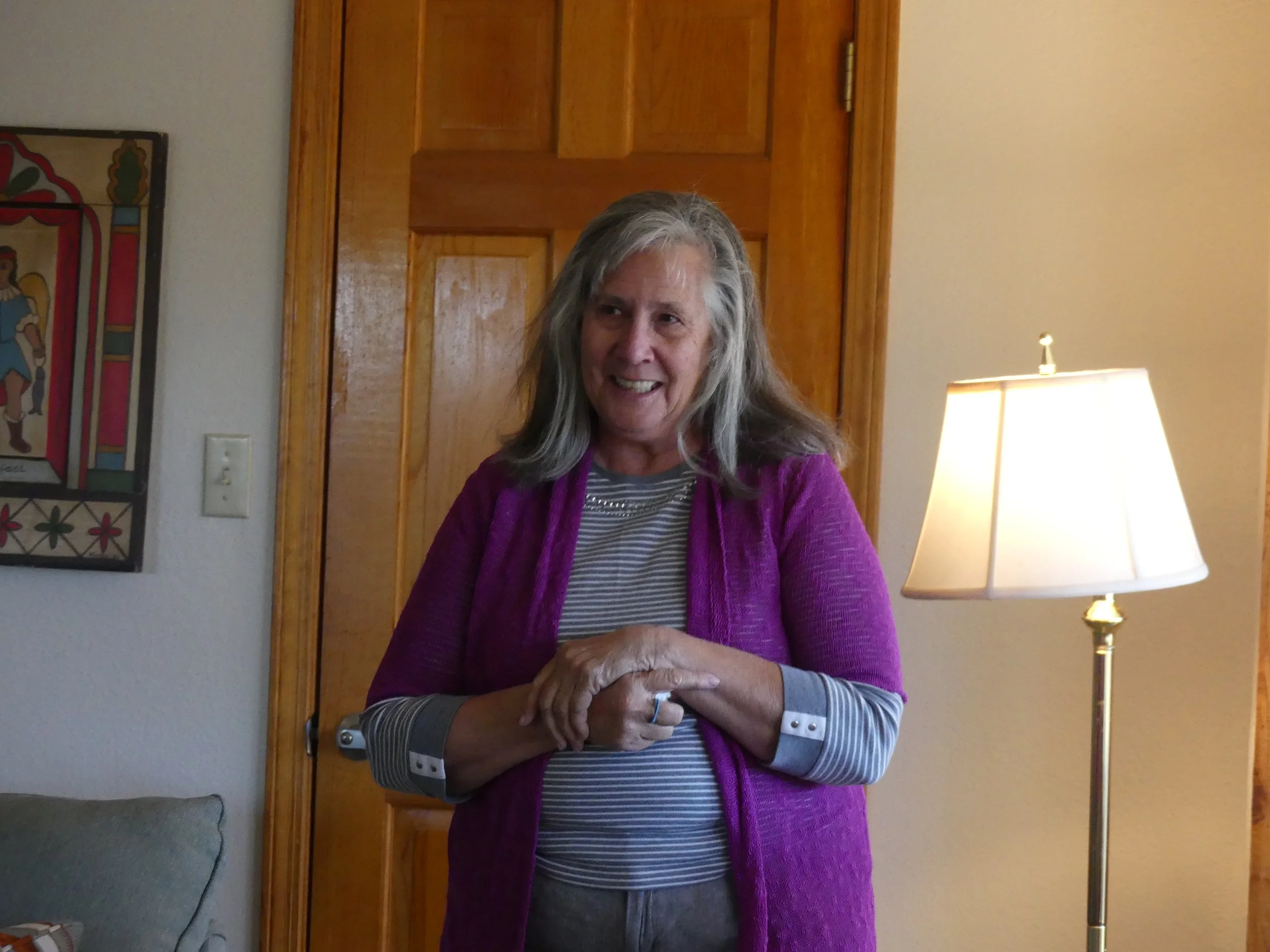 An elderly woman with gray hair smiling, wearing a black and white striped shirt and a purple cardigan, standing in a room with wooden doors, framed artwork on the wall, and a table lamp with a yellow shade.