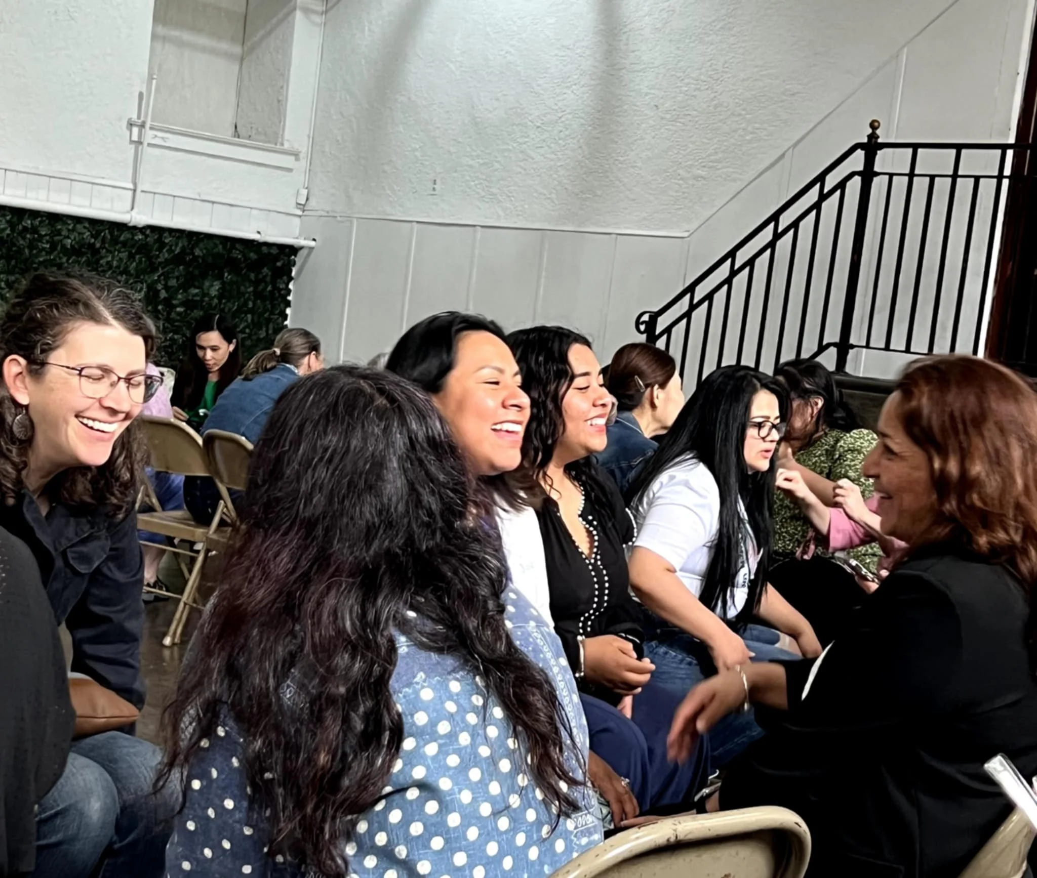 Group of people seated indoors, attentively looking at something in front of them, some smiling and engaging in conversation.