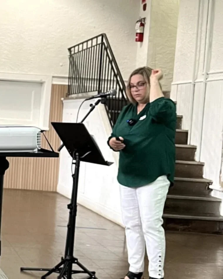 A woman in a green shirt and white pants standing at a music stand with a microphone, holding a phone, near stairs and a fire extinguisher in the background.