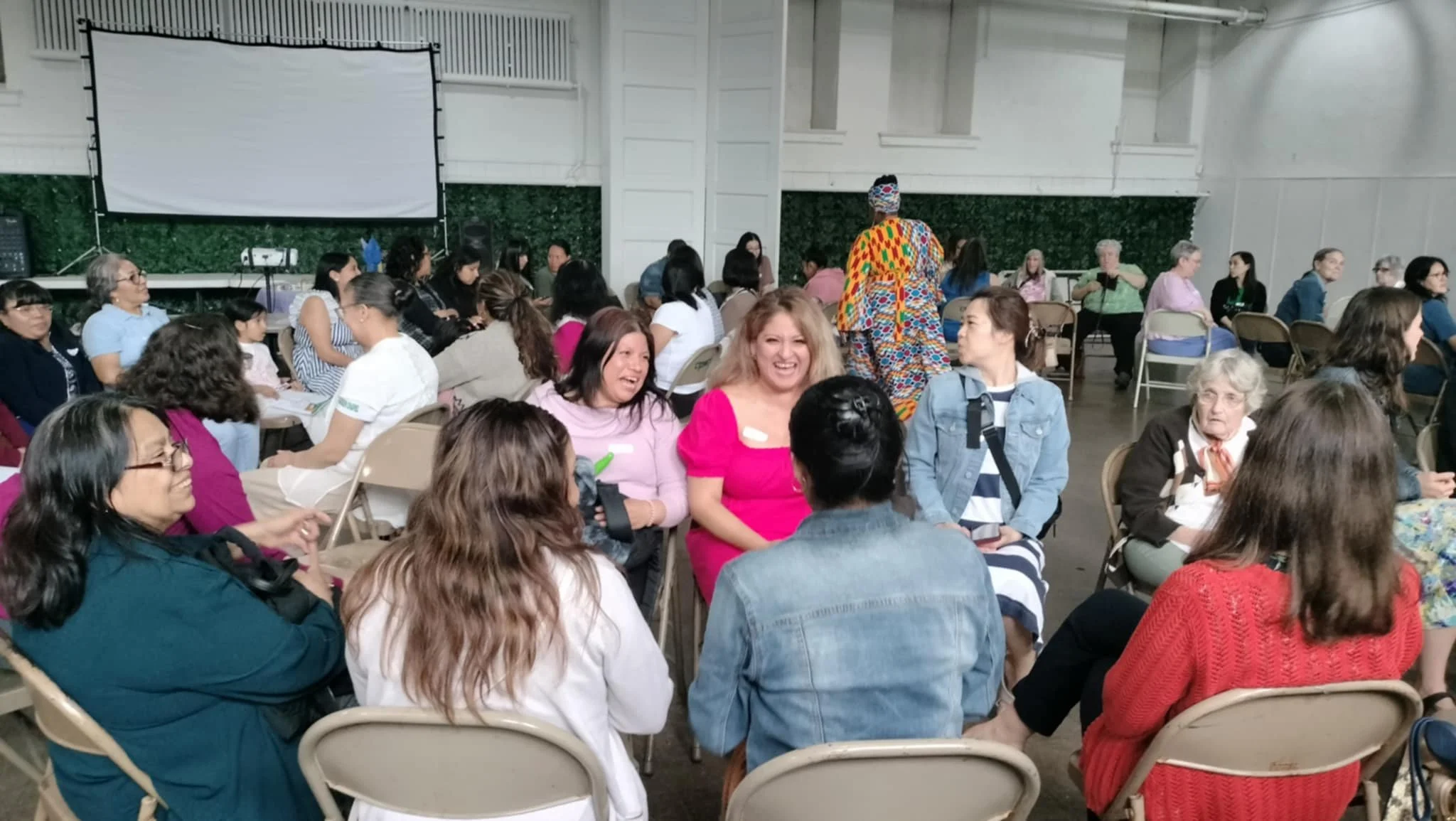 A diverse group of people sitting on beige folding chairs at a gathering, with some laughing and chatting. A woman in colorful clothing is standing among them. The setting appears to be a large indoor hall with white walls, a green hedge decoration o