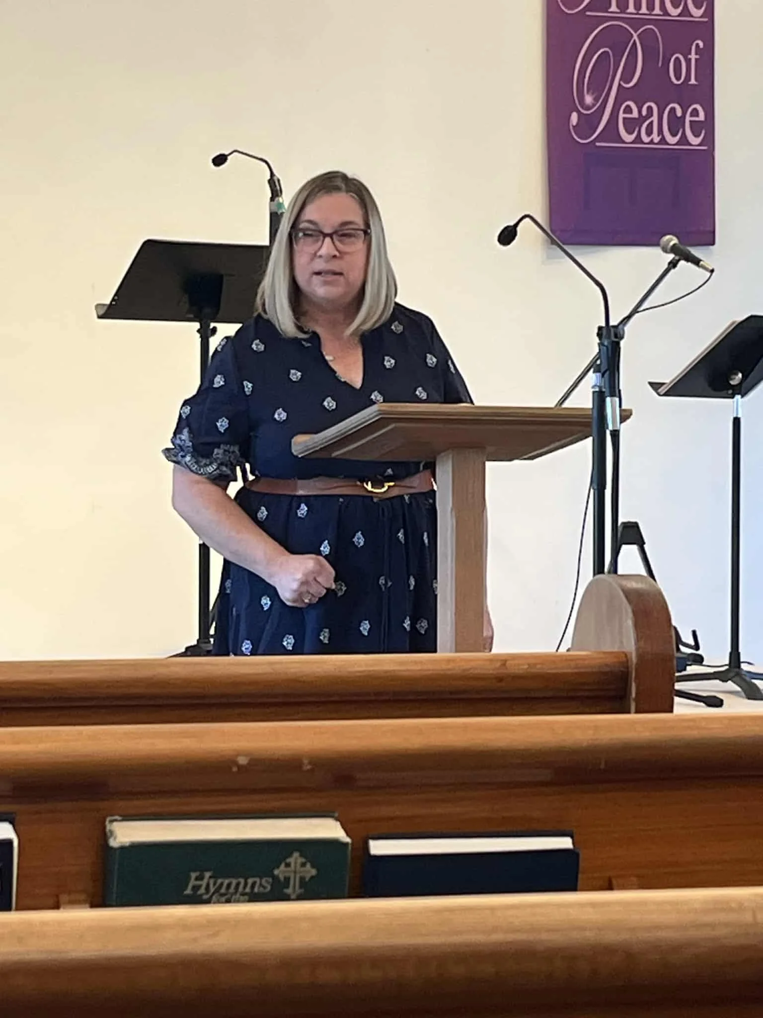 A woman with blonde hair, glasses, and a navy blue dress with white pattern standing at a pulpit in front of a church pew, in front of a sign that reads 'The Way of Peace.'