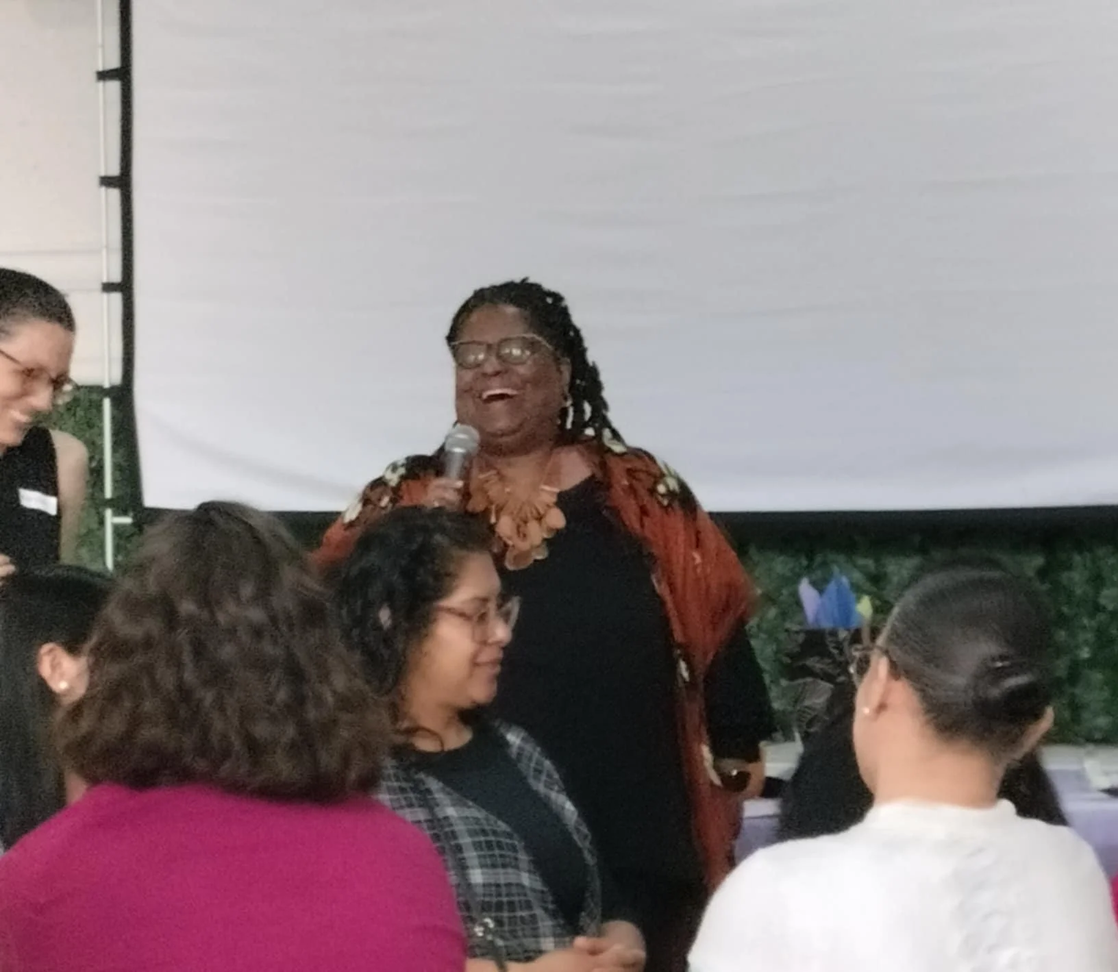 A woman with glasses and dreadlocks holding a microphone, smiling, standing in front of a group of people at an indoor event.