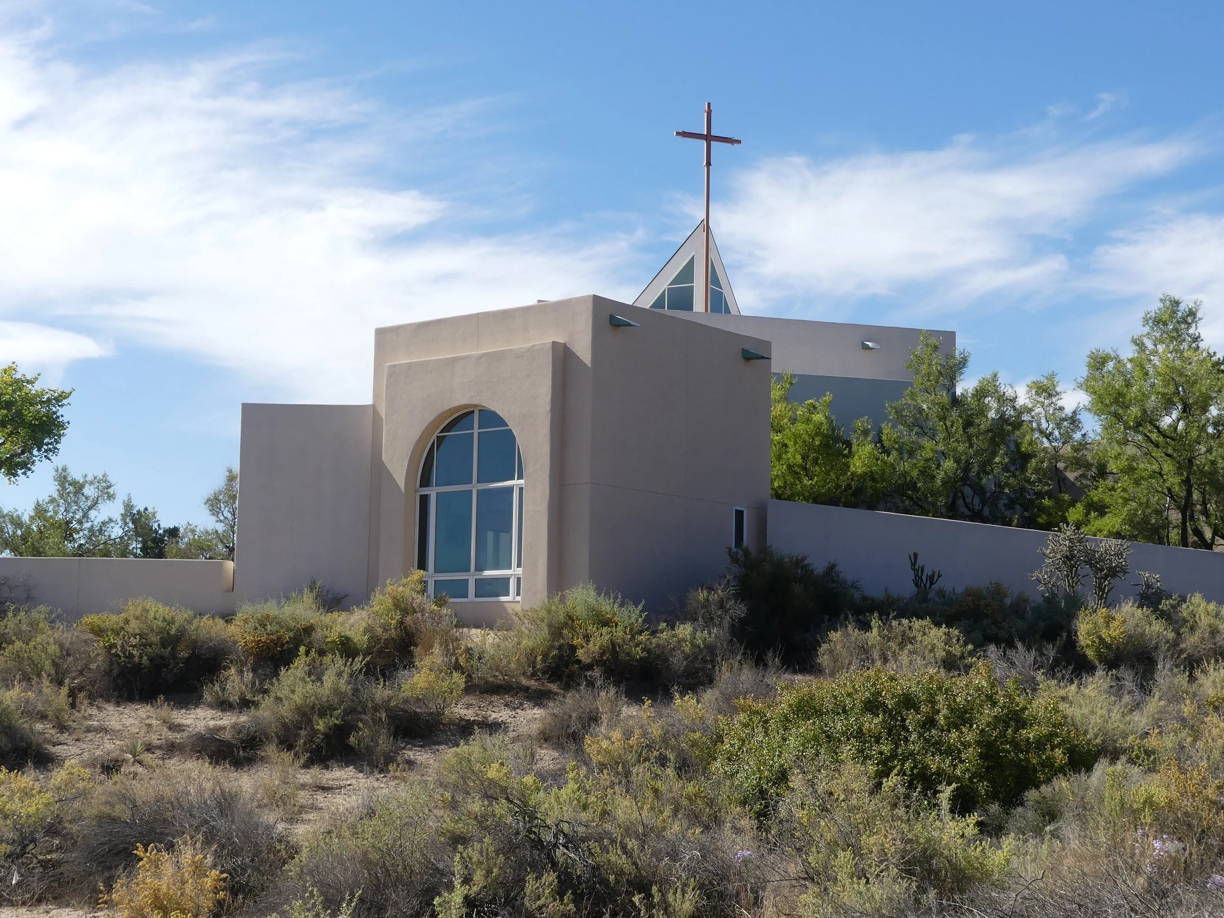 A modern church building with white walls, a large arched window, and a tall steeple topped with a cross, surrounded by trees and shrubs under a blue sky with some clouds.