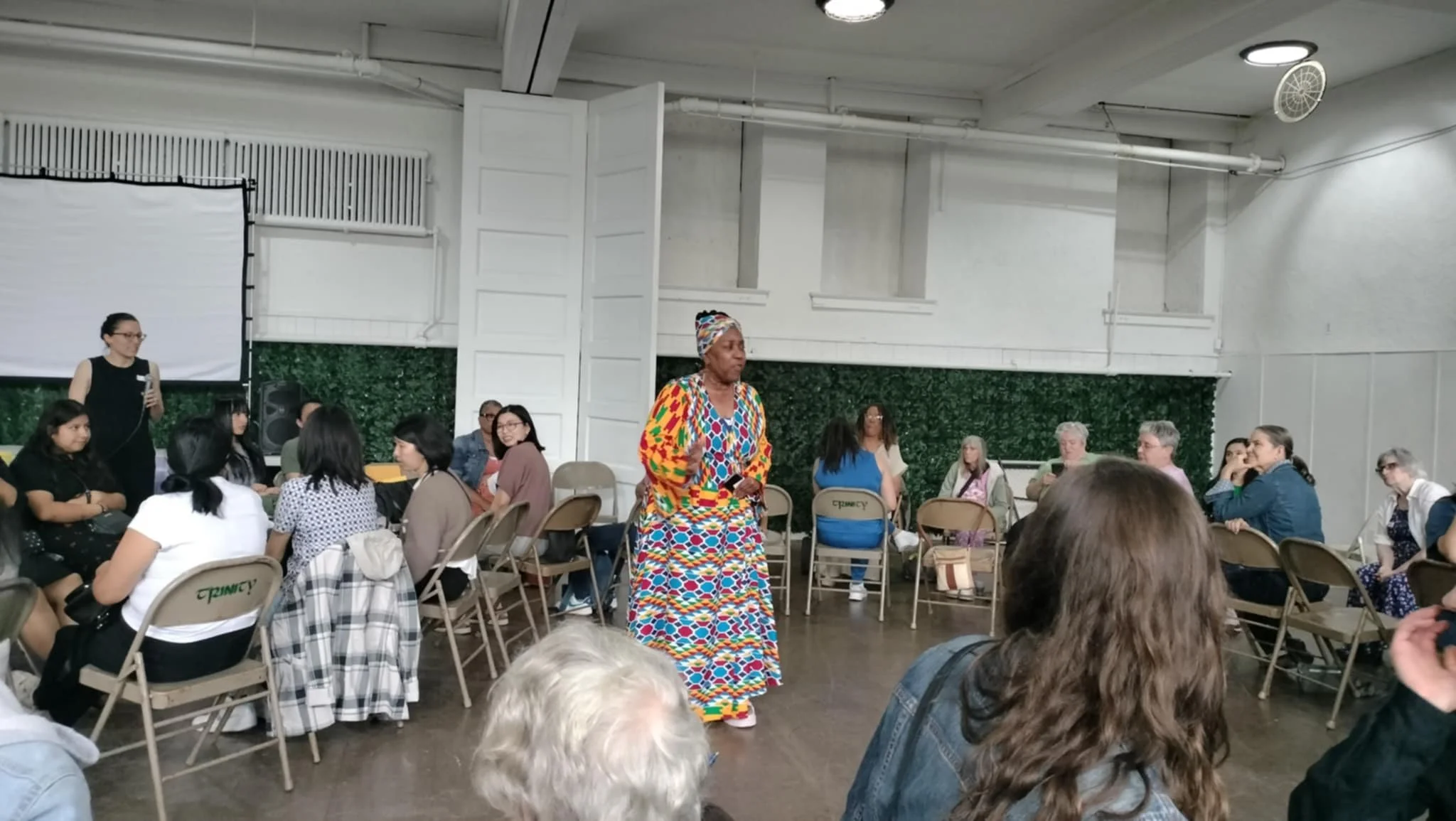 A diverse group of people attending an indoor event or meeting, with a woman in a colorful patterned dress standing in the center.