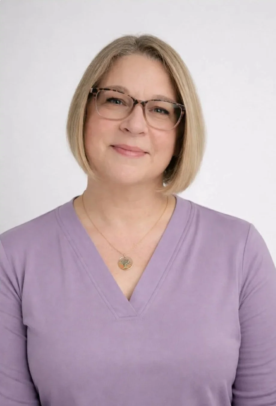 A woman with blonde hair, wearing glasses, a purple top, and a gold necklace with a tree pendant, smiling against a white background.