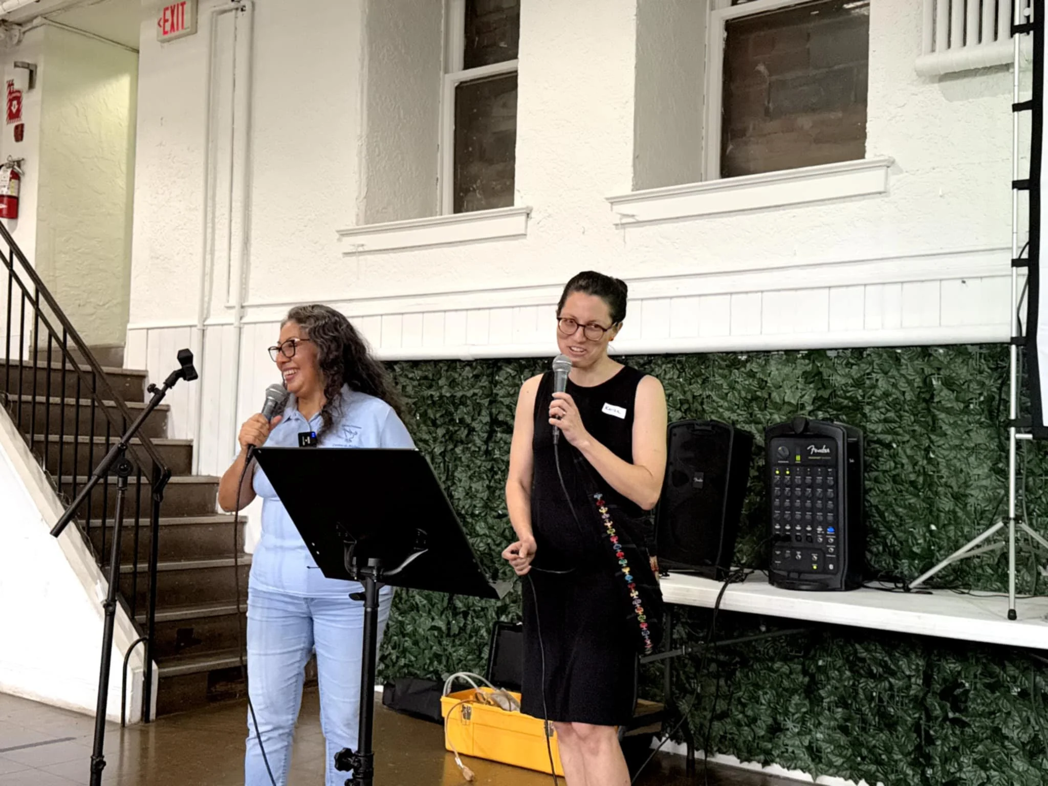 Two women standing on a stage, speaking into microphones. One woman with glasses and long hair wears a light blue top, smiling. The other woman with glasses and short hair wears a black sleeveless dress, holding a microphone and smiling. There is sou