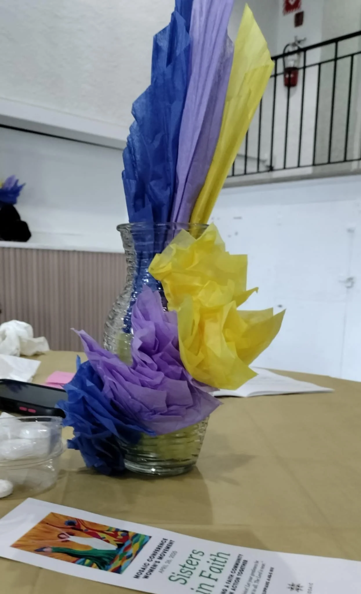 A clear glass vase with purple, yellow, and blue tissue paper decorations inside, placed on a table with papers and a flyer nearby. The background shows a white wall, a black metal railing, and a fire alarm.