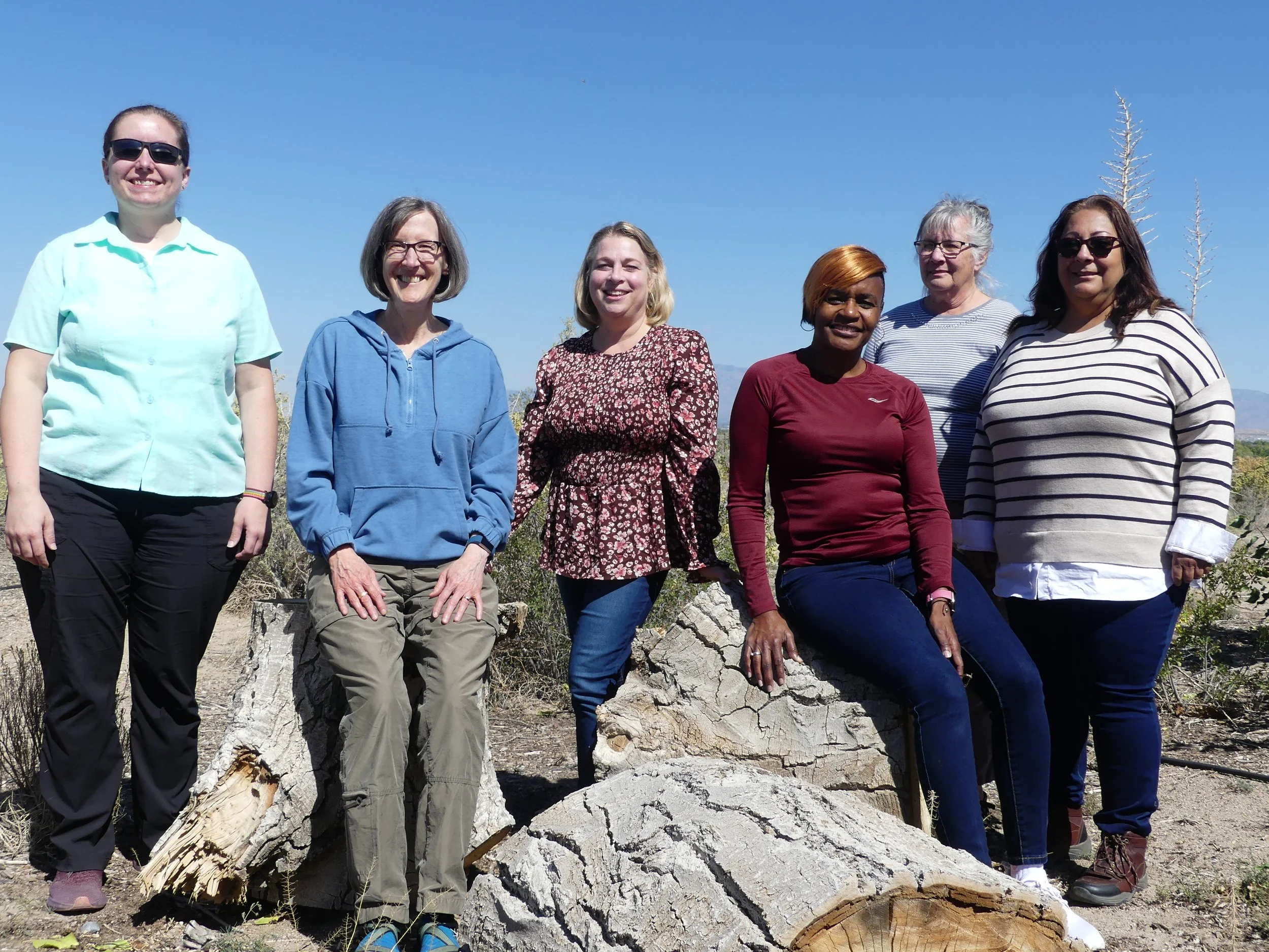 A group of seven women standing outdoors on a sunny day, smiling, with a large fallen tree trunk in front of them and leafless trees in the background.