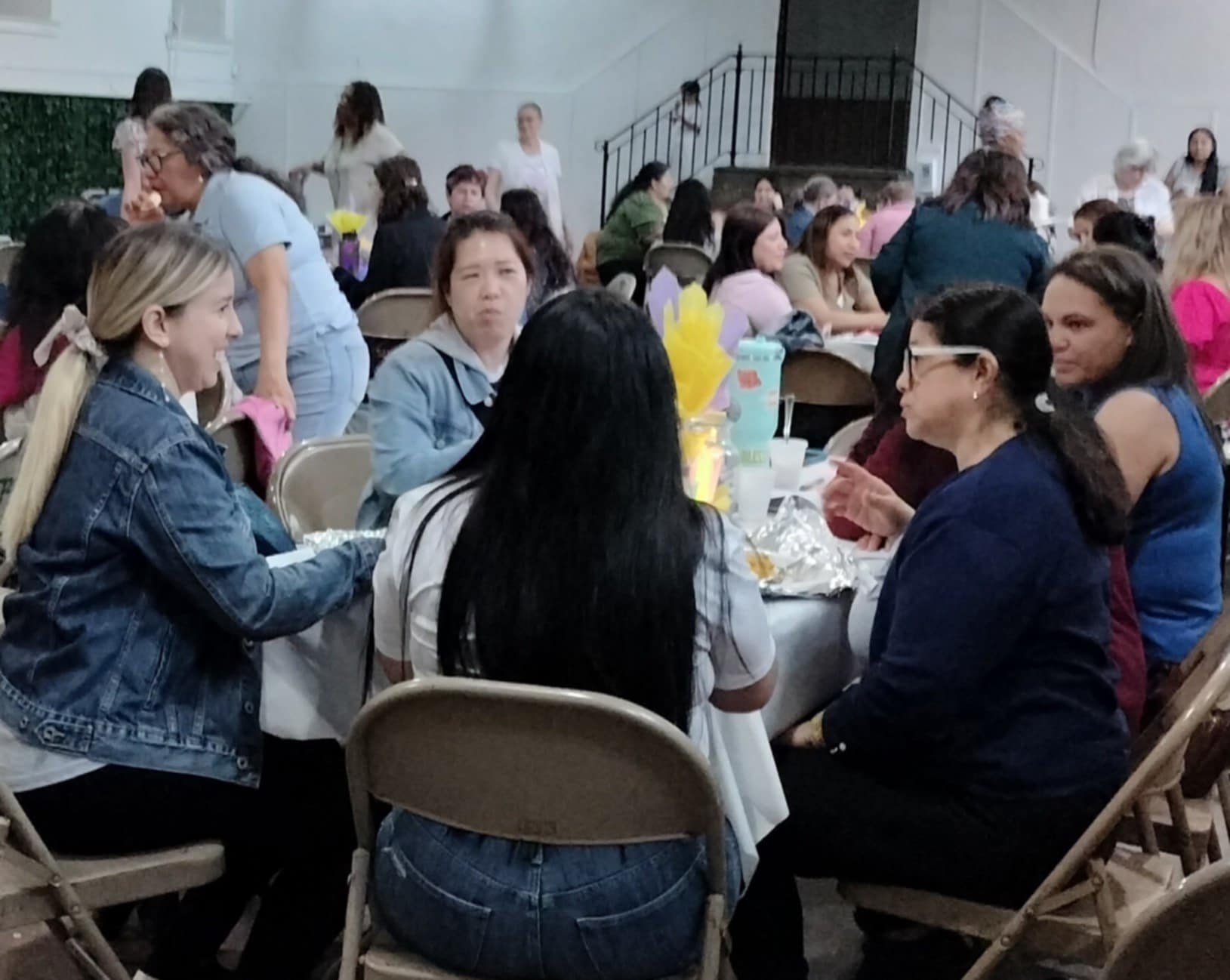 A group of women are sitting around a table at a social gathering or event, engaged in conversation, with some standing or walking in the background.