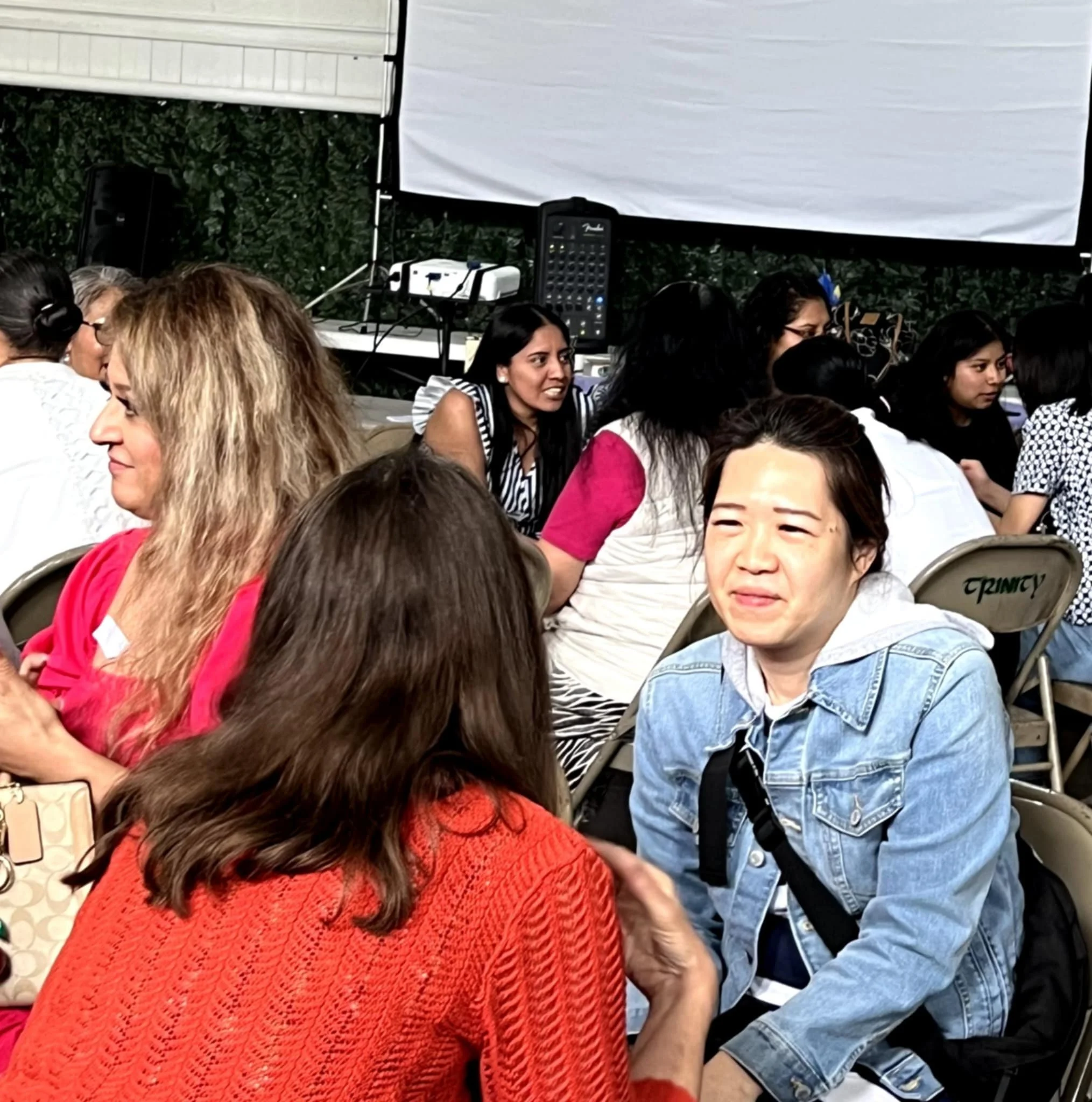 People sitting closely together at an outdoor event with a speaker and a screen in the background.