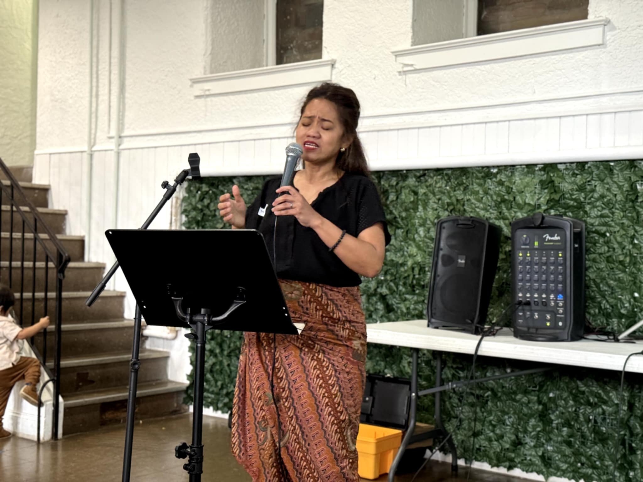 A woman singing into a microphone at an indoor event. She is dressed in a black top and a patterned skirt, standing behind a music stand, with speakers and other audio equipment nearby. There is a green leafy backdrop behind her and a staircase to th
