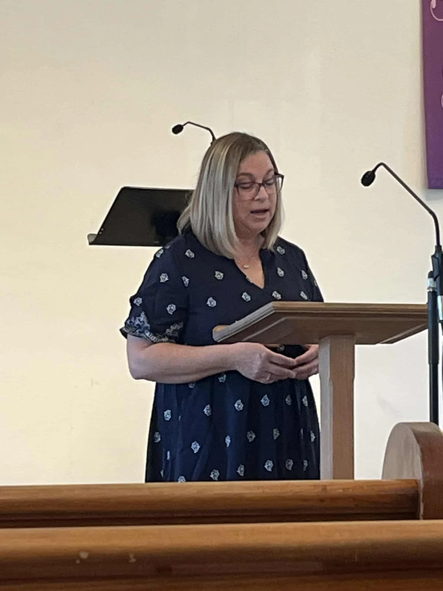 A woman with blonde hair, glasses, wearing a dark dress with a light pattern, stands at a podium with a microphone, reading from a paper during a speech or presentation in a room with white walls.