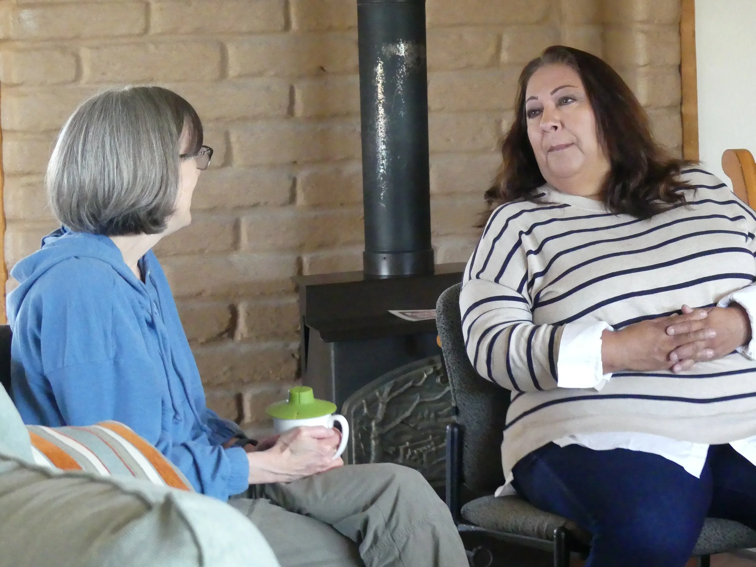Two women engaged in conversation, one with gray hair and glasses wearing a blue jacket, and the other with dark brown hair wearing a striped shirt, sitting in a room with a brick wall and a black stove in the background. The woman in the striped shi