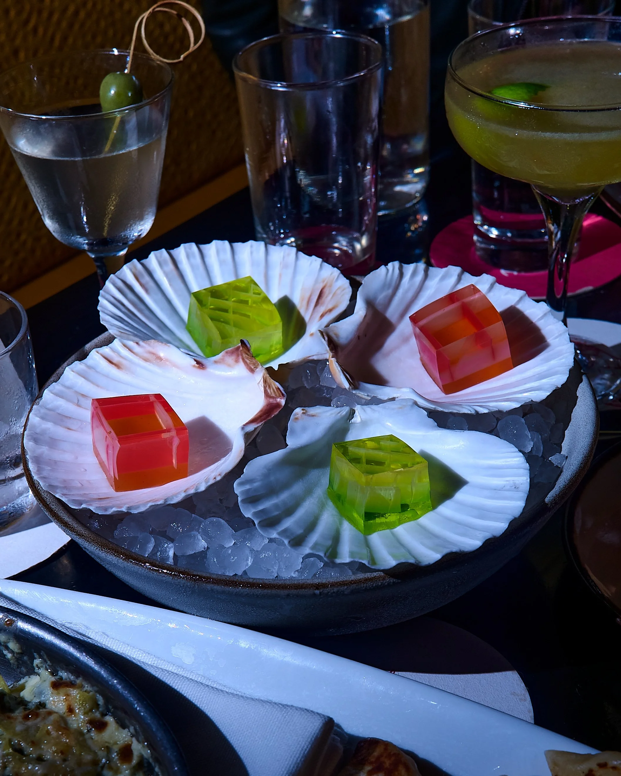 A tray of four multicolored Jello shots presented in white oyster shells, set on a table with cocktails in stemmed glassware and empty water glasses in the background.