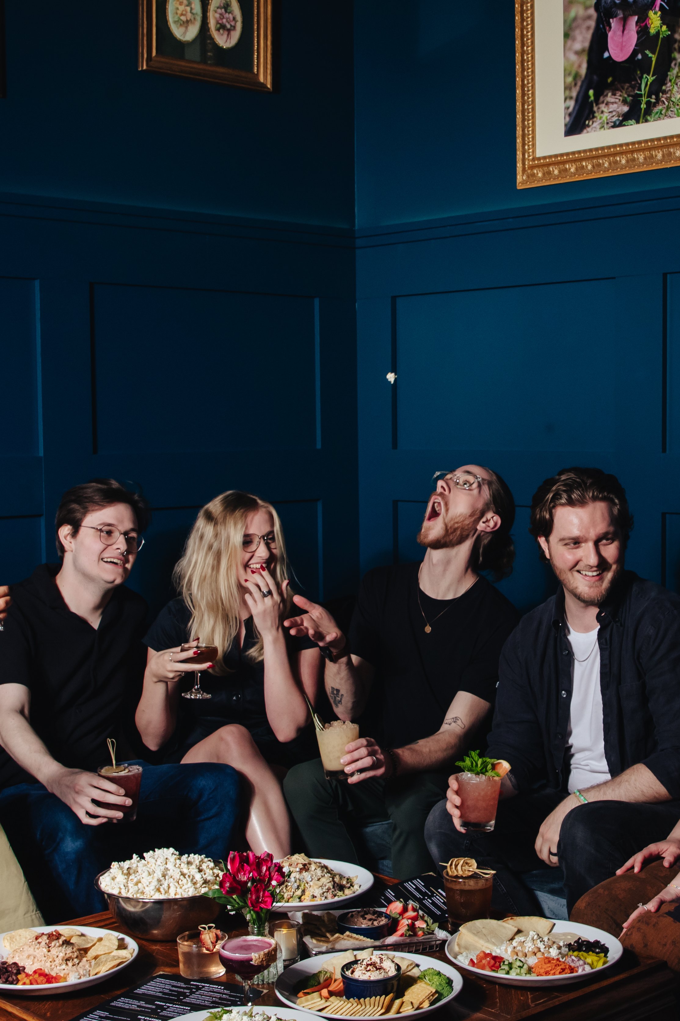 Four young adults sitting on a couch at a party, laughing and enjoying drinks, with a table of food and snacks in front of them.