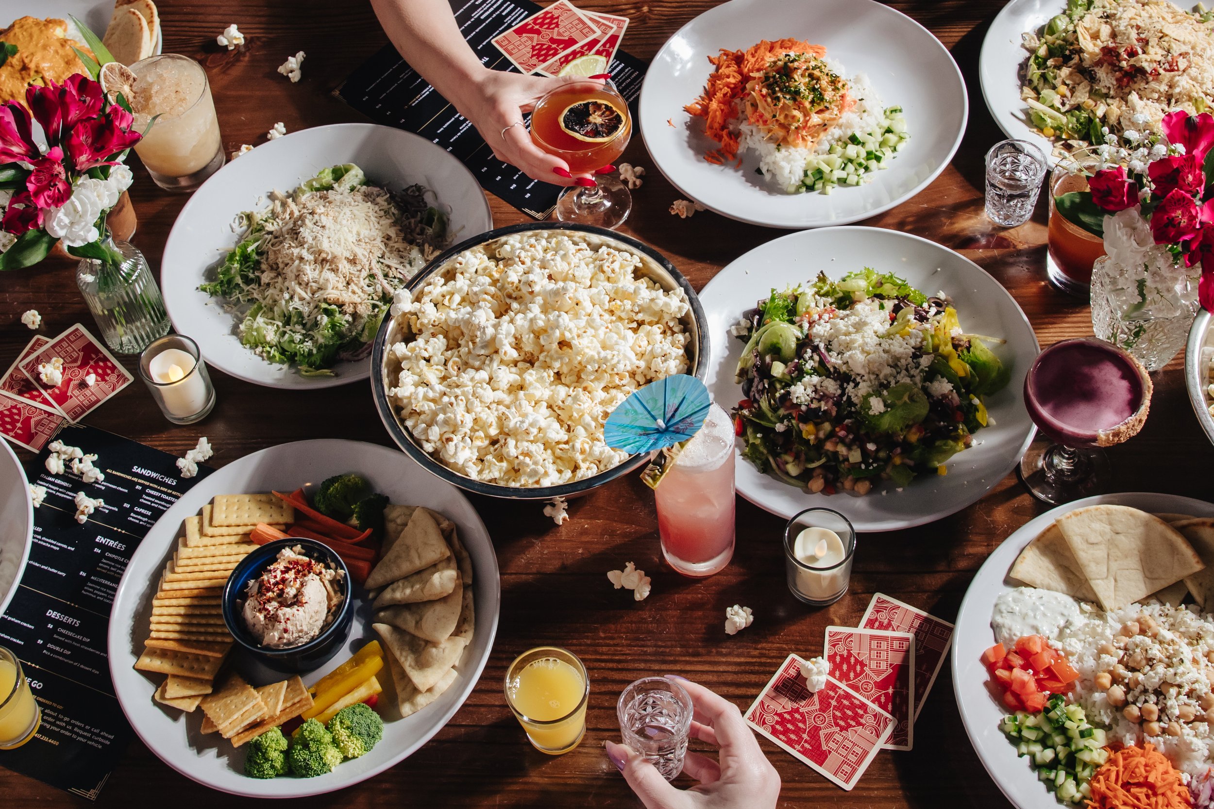A festive dinner table with various dishes including popcorn, salads, and chips with dip, decorated with flowers, candles, and playing cards, with people holding drinks.