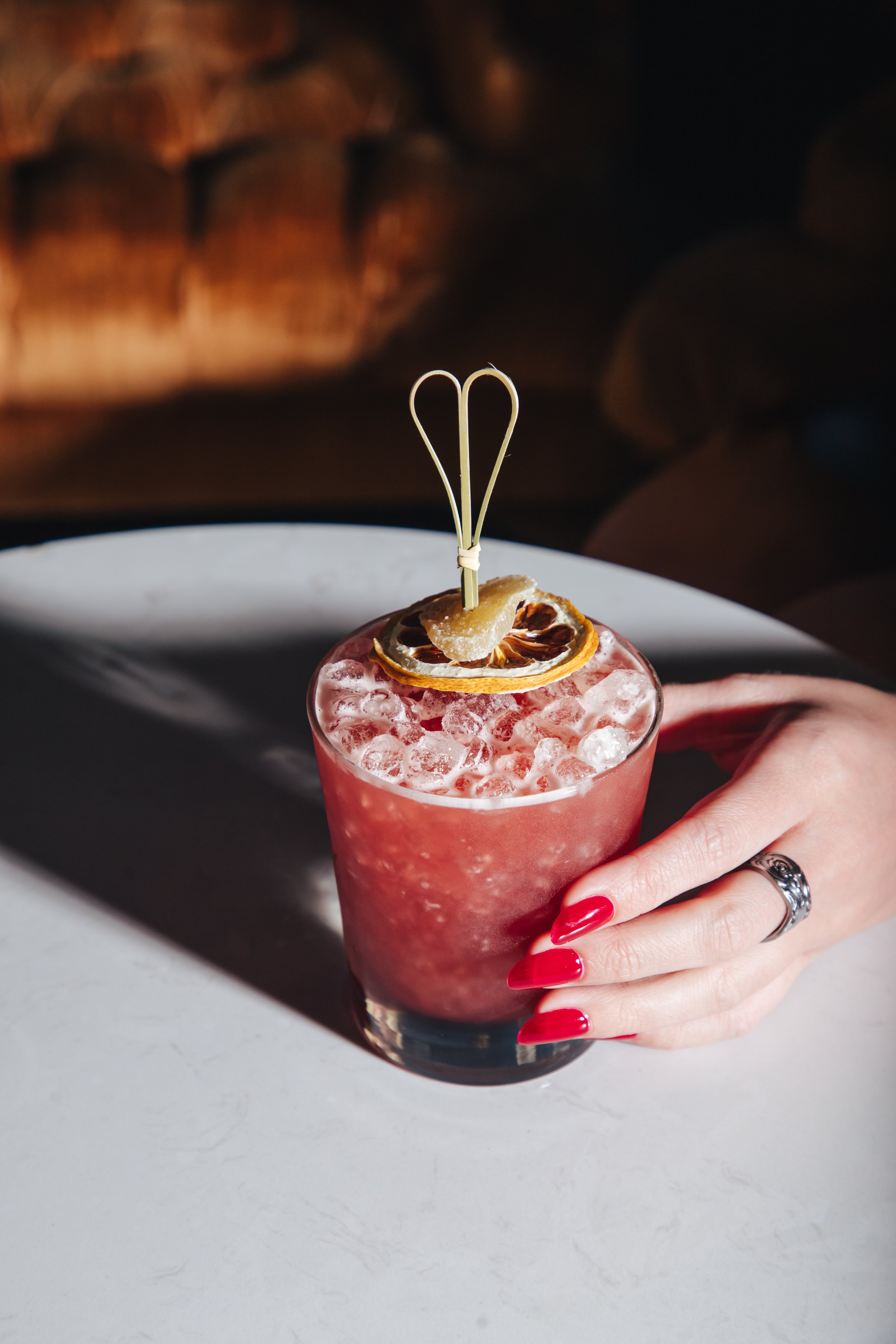 A person with red nail polish and a silver ring on their finger holds a pink cocktail in a clear glass, garnished with dried lemon slices and a decorative stir stick. The cocktail is filled with ice and is on a white surface with a blurred, warmly li