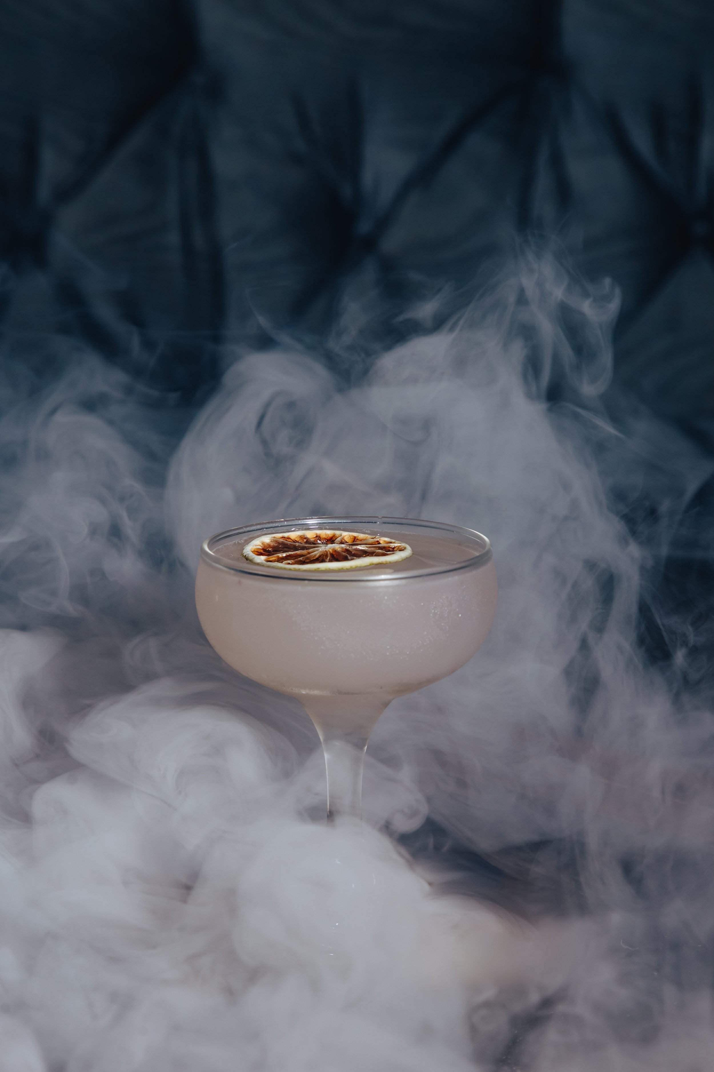 A cocktail in a coupe glass garnished with a dried lime slice, surrounded by swirling smoke or vapor.