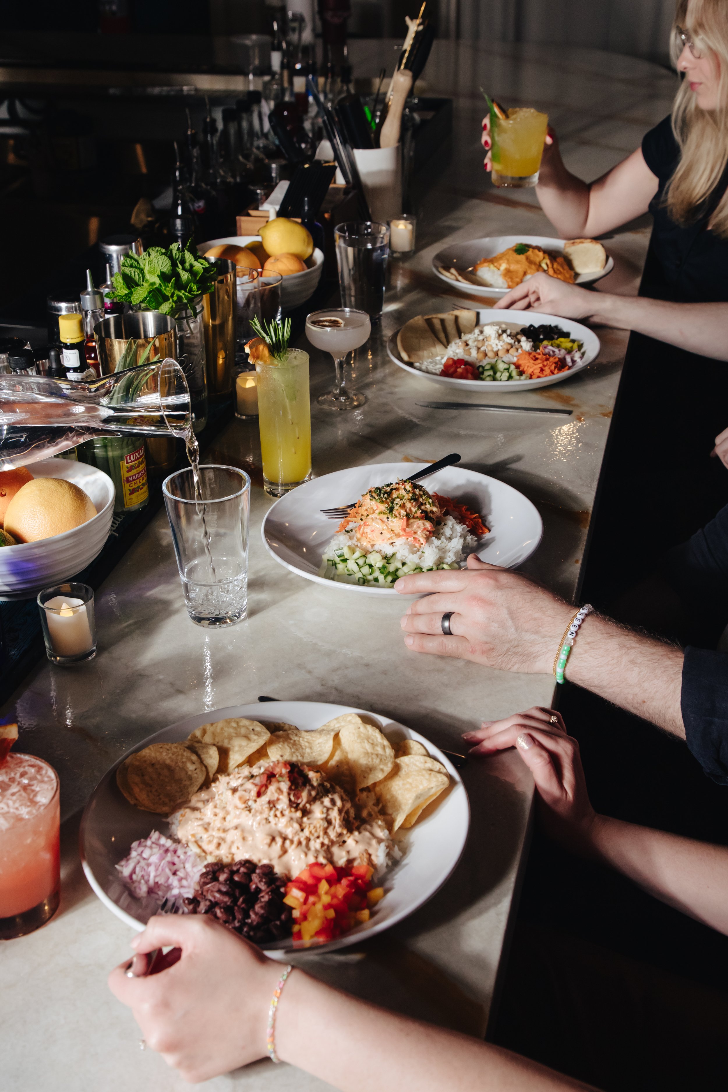 A bar or restaurant counter with various dishes of food and drinks, including salads, a plate of chips with dip, cocktails, and fresh fruits, with people serving and enjoying their meals.
