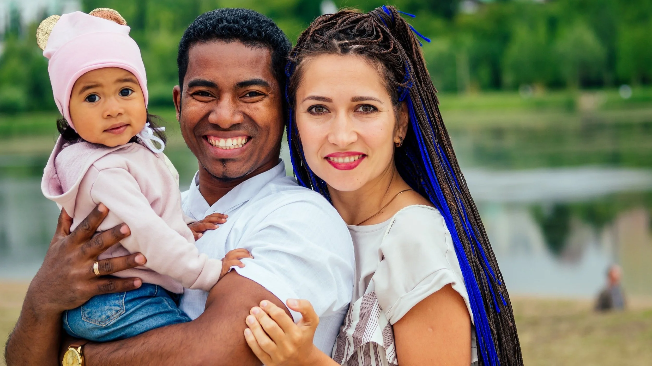 A smiling couple holding a baby near a body of water, with greenery in the background. The baby is wearing a pink hat.