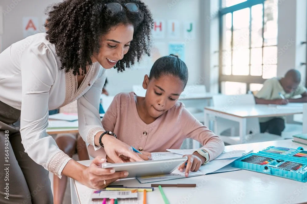 Teacher assisting a student with a tablet in a classroom setting, surrounded by school supplies.