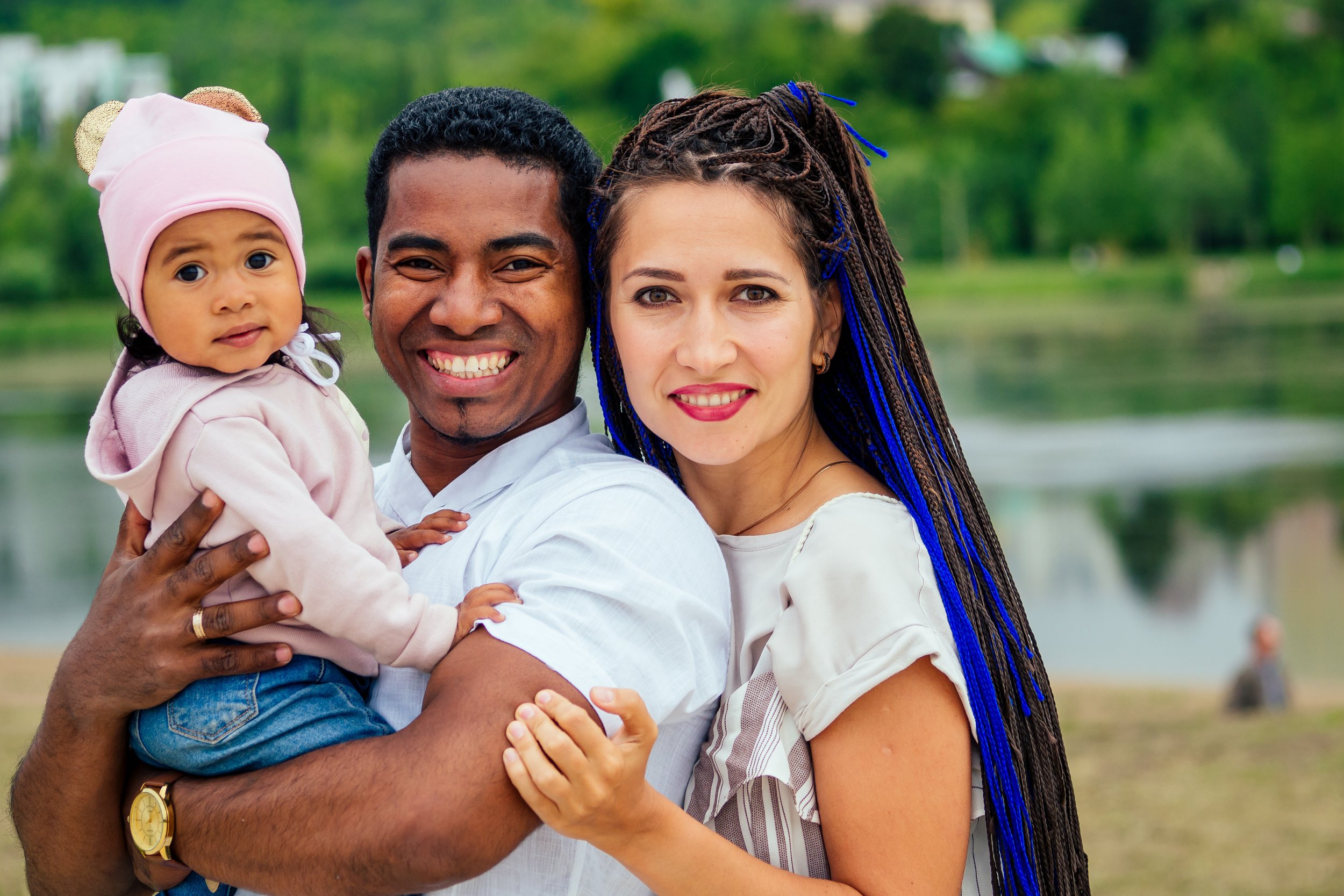A happy couple with a baby posing outdoors near a lake. The man is holding the baby, and the woman is standing beside them. The baby is wearing a pink hat, and the woman has long braids with blue highlights. The background features trees and water.
