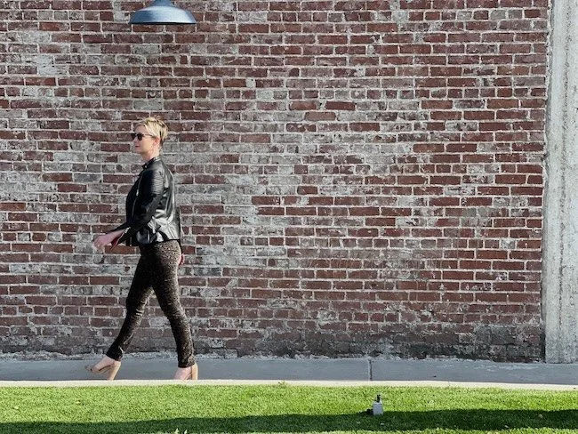 Ashlee Lynn Espinosa walking along a sunlit brick wall in a leather jacket, embodying freedom, flow, and modern creative living.
