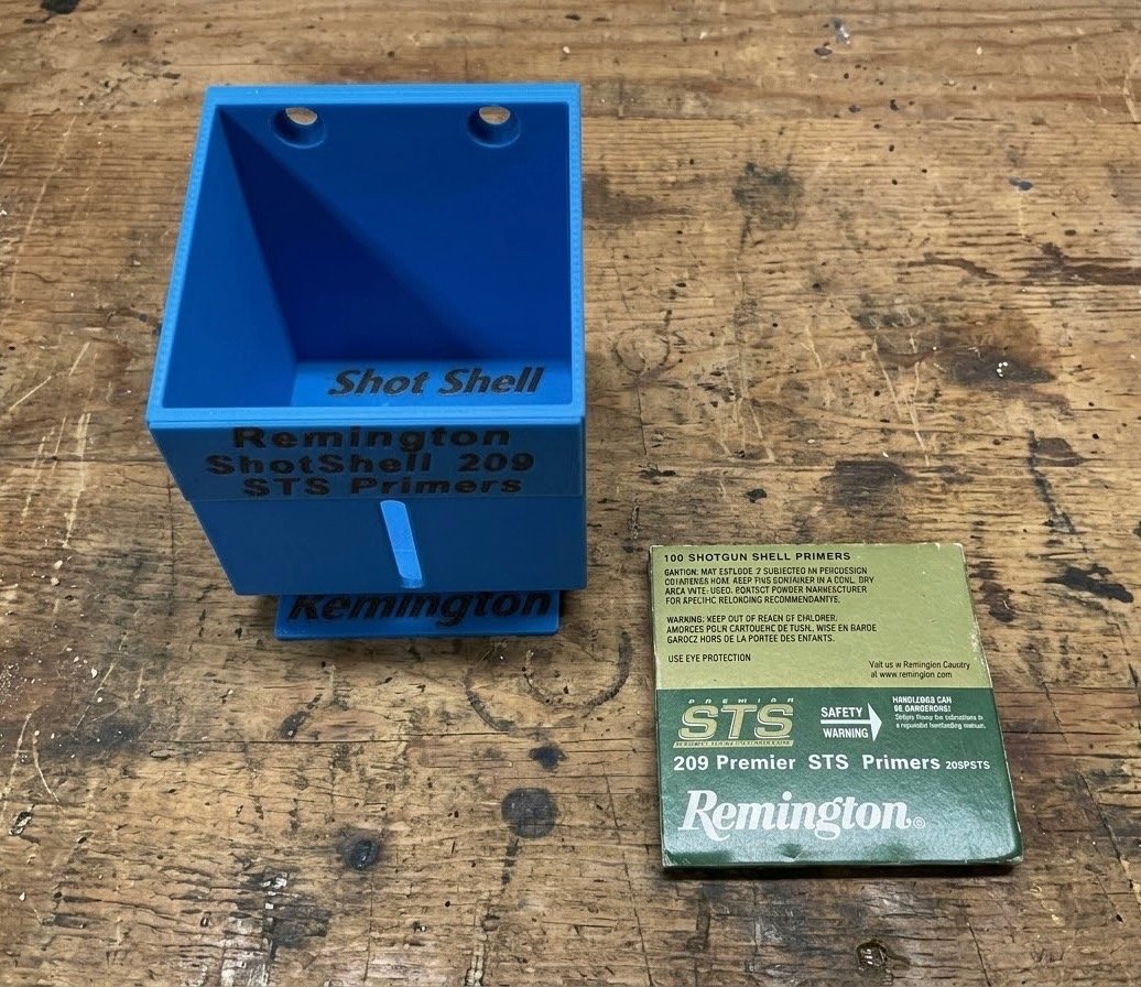 A blue Remington shot shell primer holder sitting on wooden surface next to primer pack.