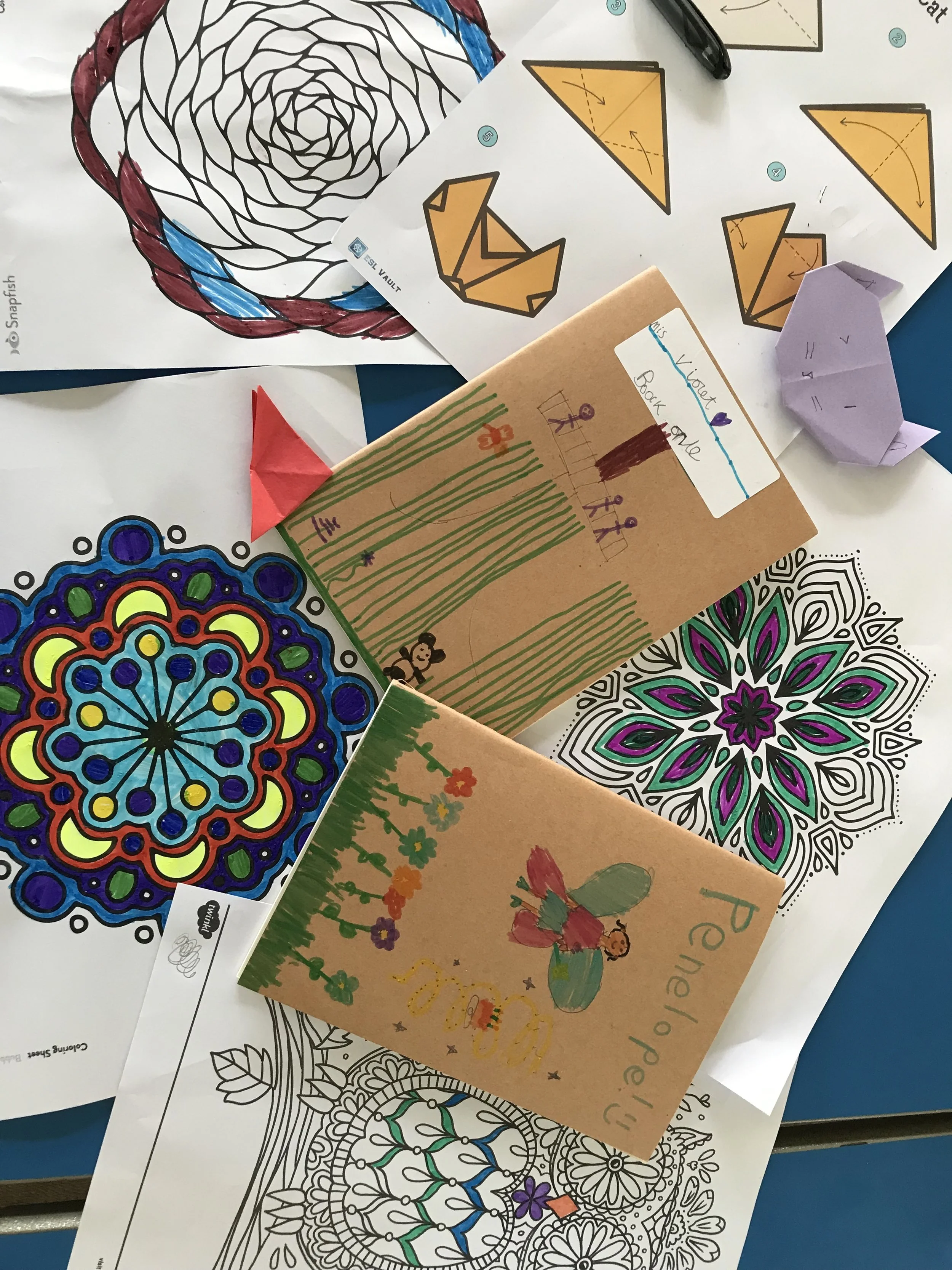 Various coloring pages and drawings on paper, including mandala patterns, origami instructions, and a child's drawing with a butterfly and flowers.