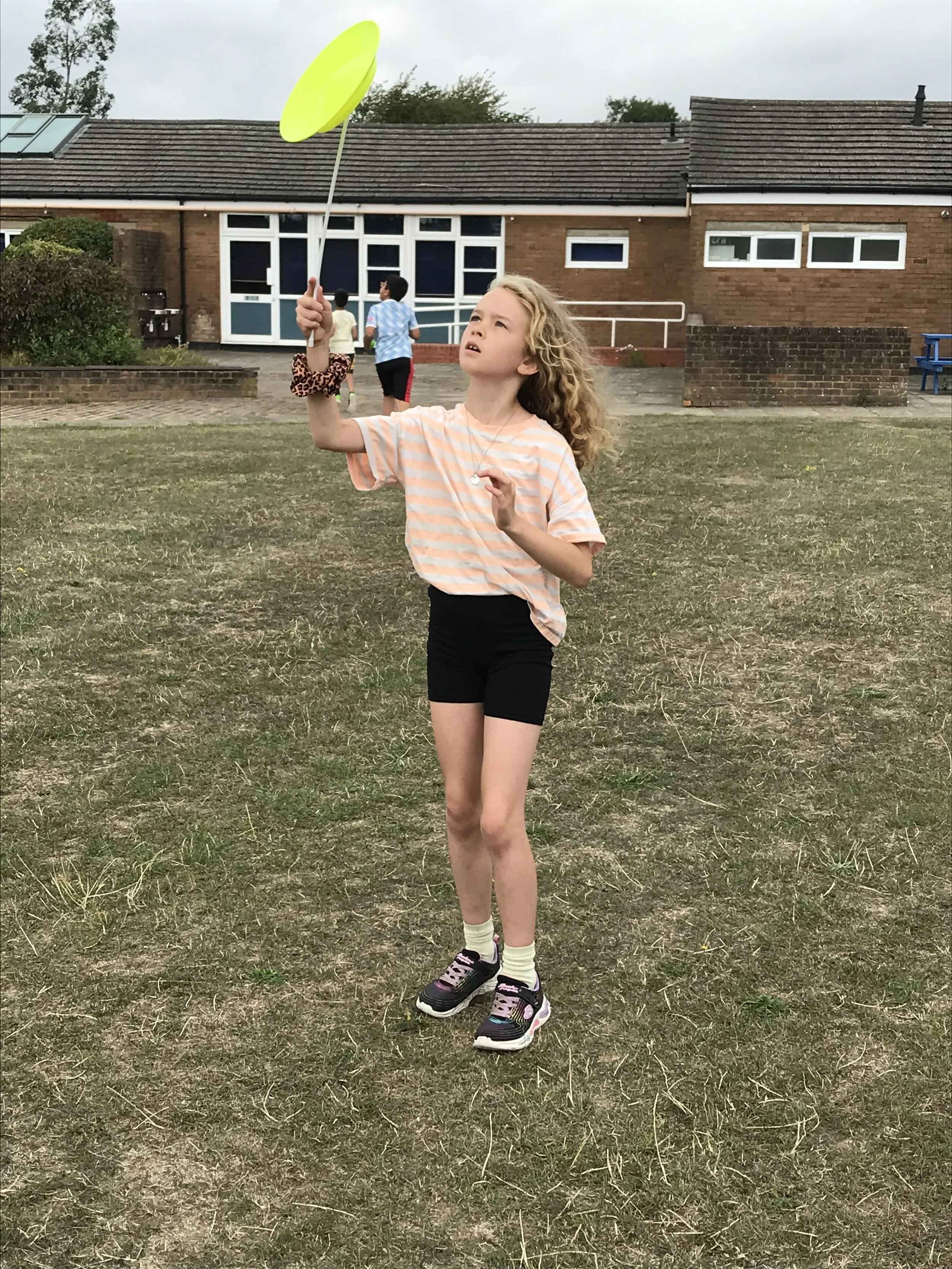 A young girl with curly blonde hair playing with a yellow spinning disc outdoors on a grassy field, with a brick school building and children in the background.