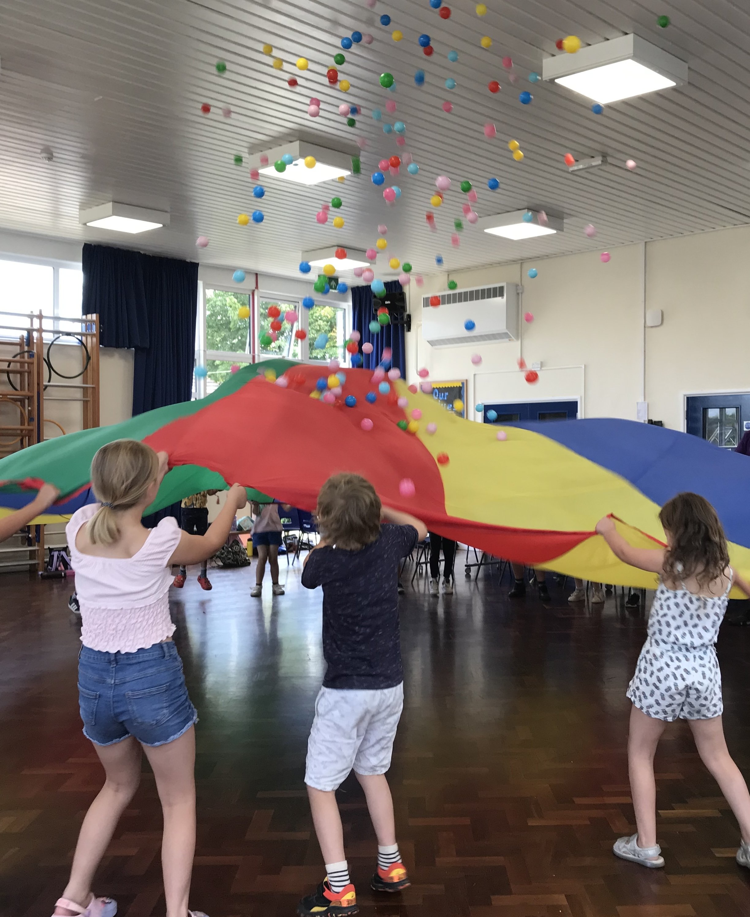 Children playing with a colorful parachute indoors, with some balls being thrown into the air.