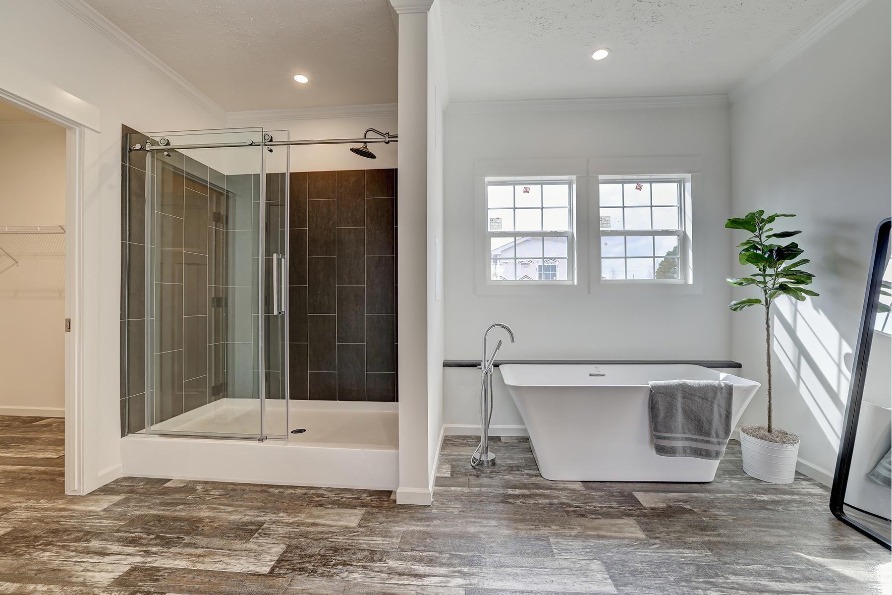 Modern bathroom with a glass-enclosed shower, freestanding bathtub with towel, large windows, potted plant, and wooden flooring.