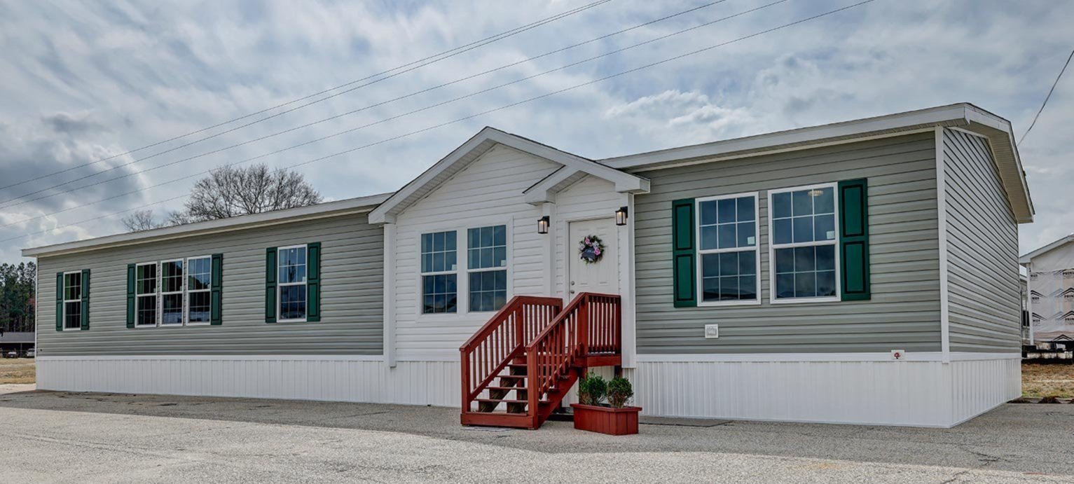 Single-story manufactured home with green siding, white trim, and green shutters, featuring a red front staircase, set under a partly cloudy sky.