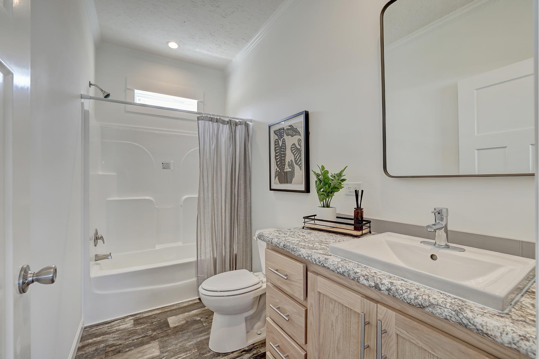 Modern bathroom with wood vanity, granite countertop, square mirror, bathtub with shower curtain, toilet, framed artwork, and potted plant decoration.