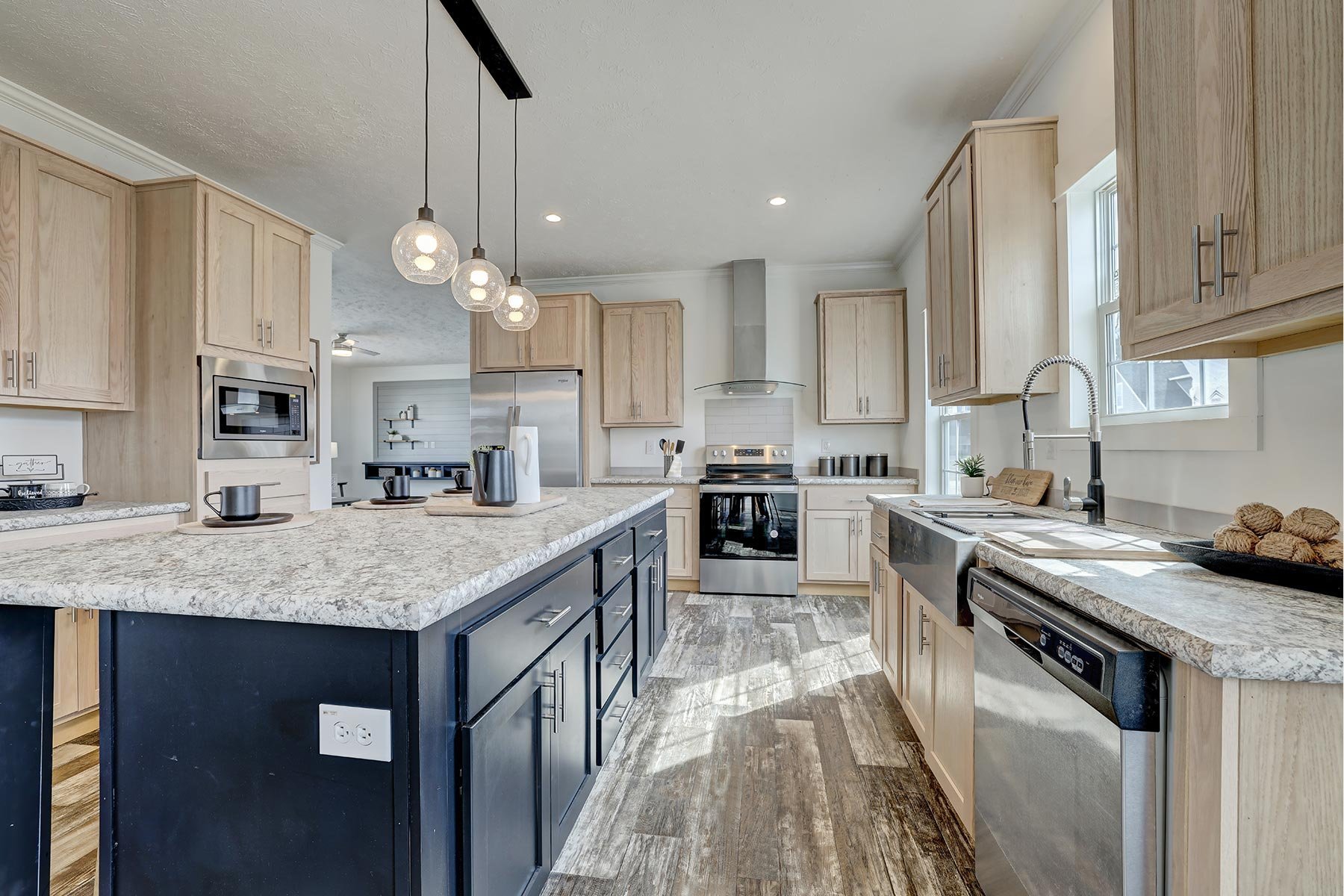 Modern kitchen with wooden cabinets, granite countertops, and stainless steel appliances, including a refrigerator, oven, and dishwasher. A large island with pendant lighting and barstools is centrally located. Light gray flooring and natural light e