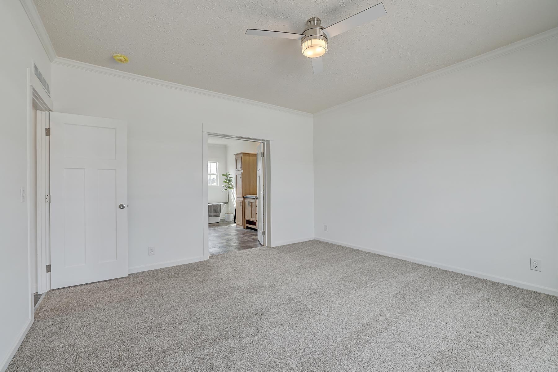 Empty room with carpet flooring, ceiling fan, closed white door, and adjacent room visible through open door with wooden cabinet and window.