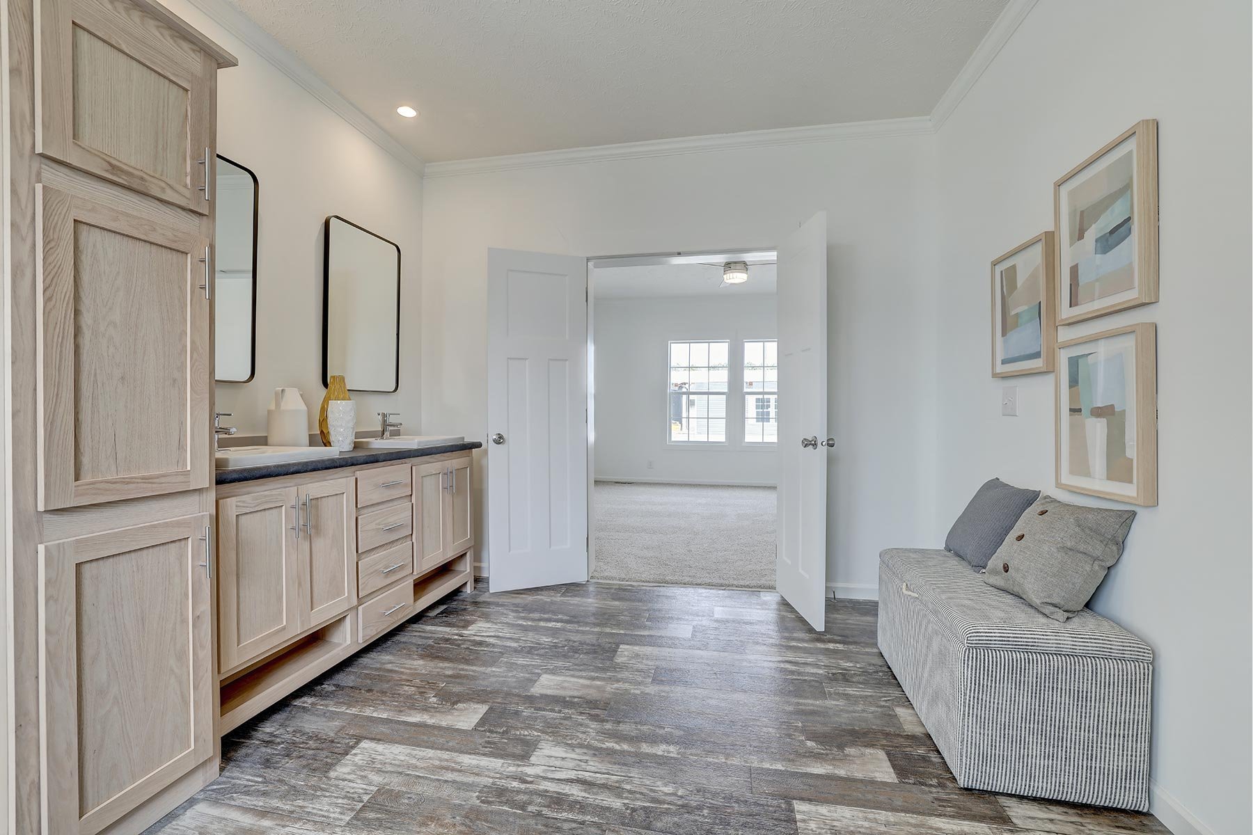 Modern bathroom interior with double vanity, wood cabinets, two mirrors, abstract wall art, striped bench, and open doors leading to a bright room with carpet.